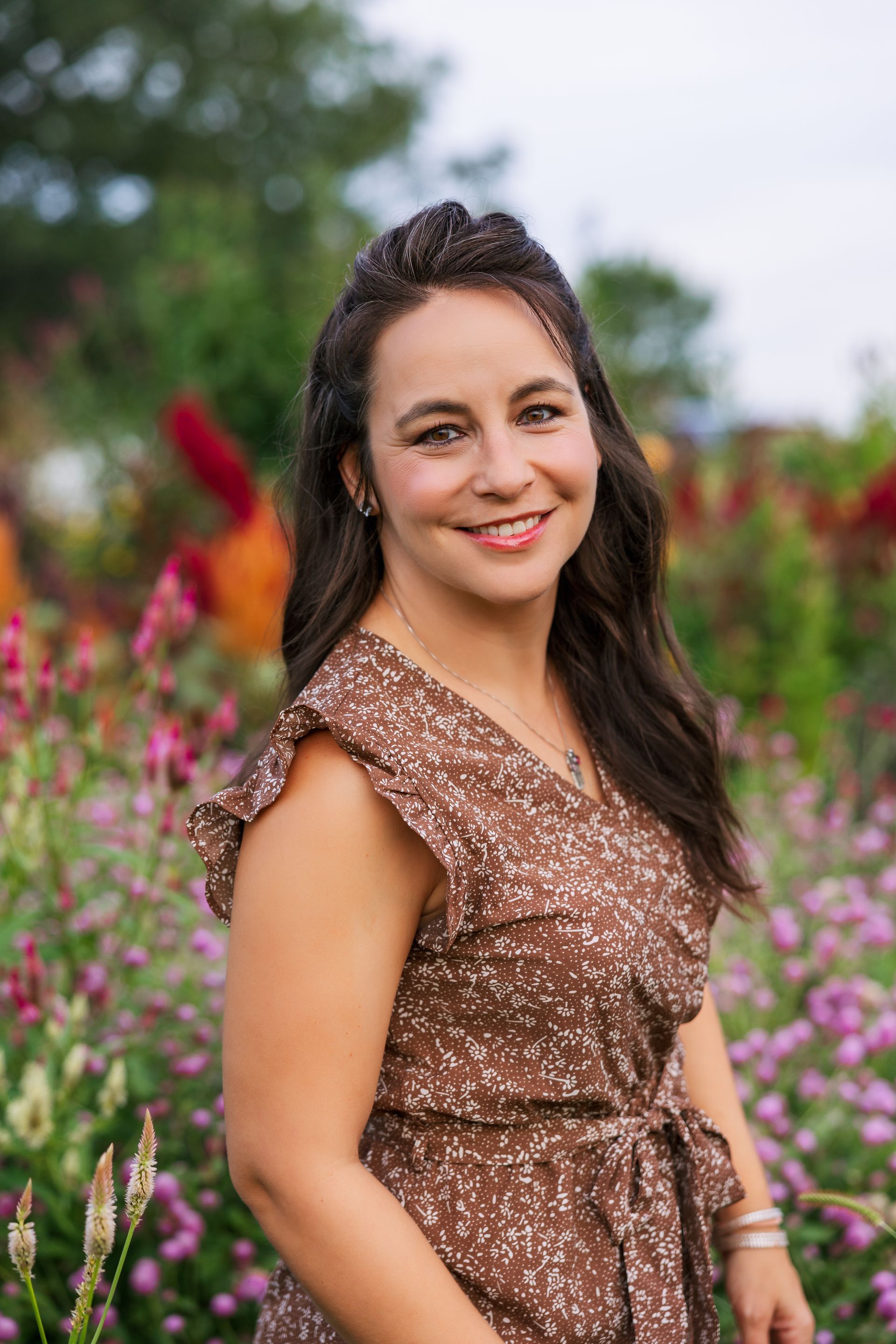Woman with brown hair, smiling, in a brown floral dress outdoors near colorful flowers.