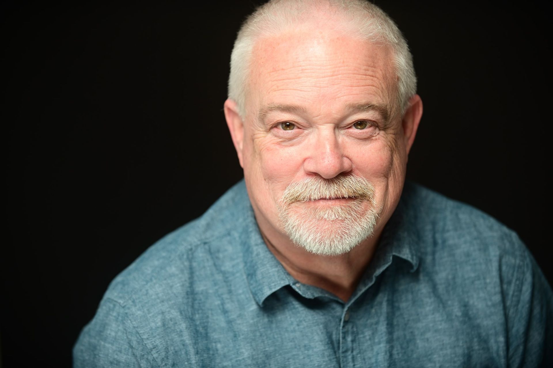 Smiling older man with a white goatee and light blue shirt against a black background.