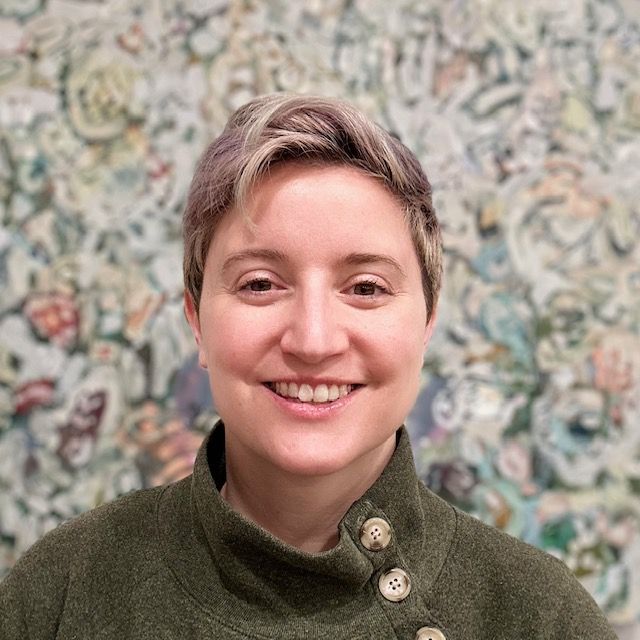 Woman with short, purple-grey hair, smiling, wearing a green sweater. In front of a colorful, abstract background.