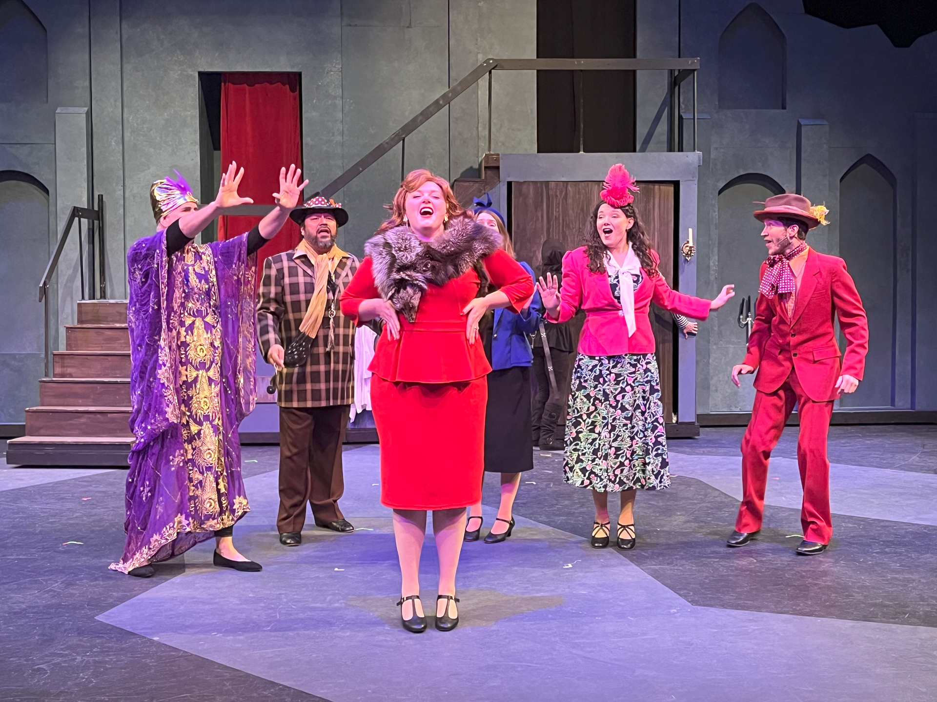 A theater cast in bright outfits, laughing and gesturing on stage.