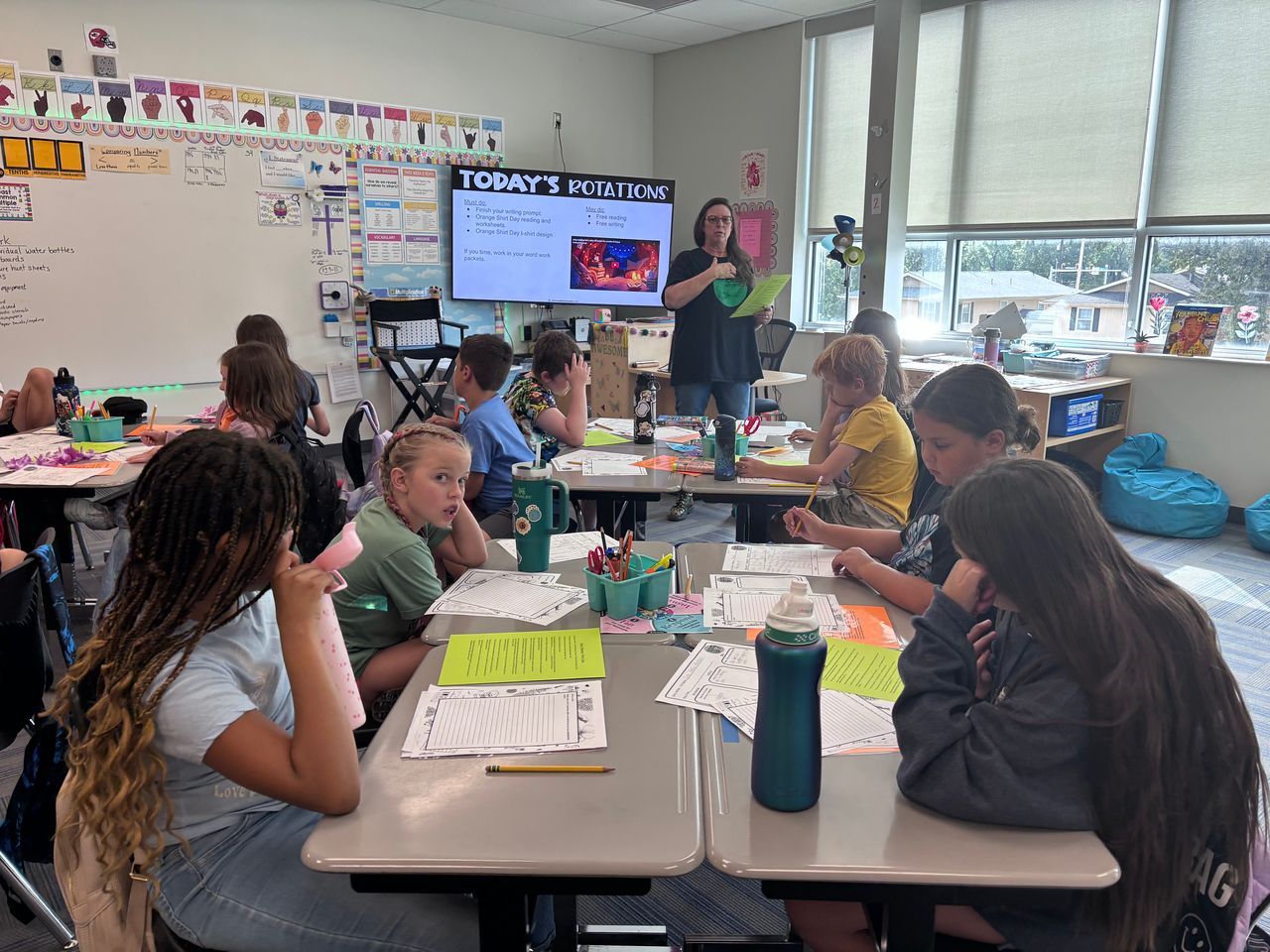 Classroom scene: students at desks, teacher at screen.