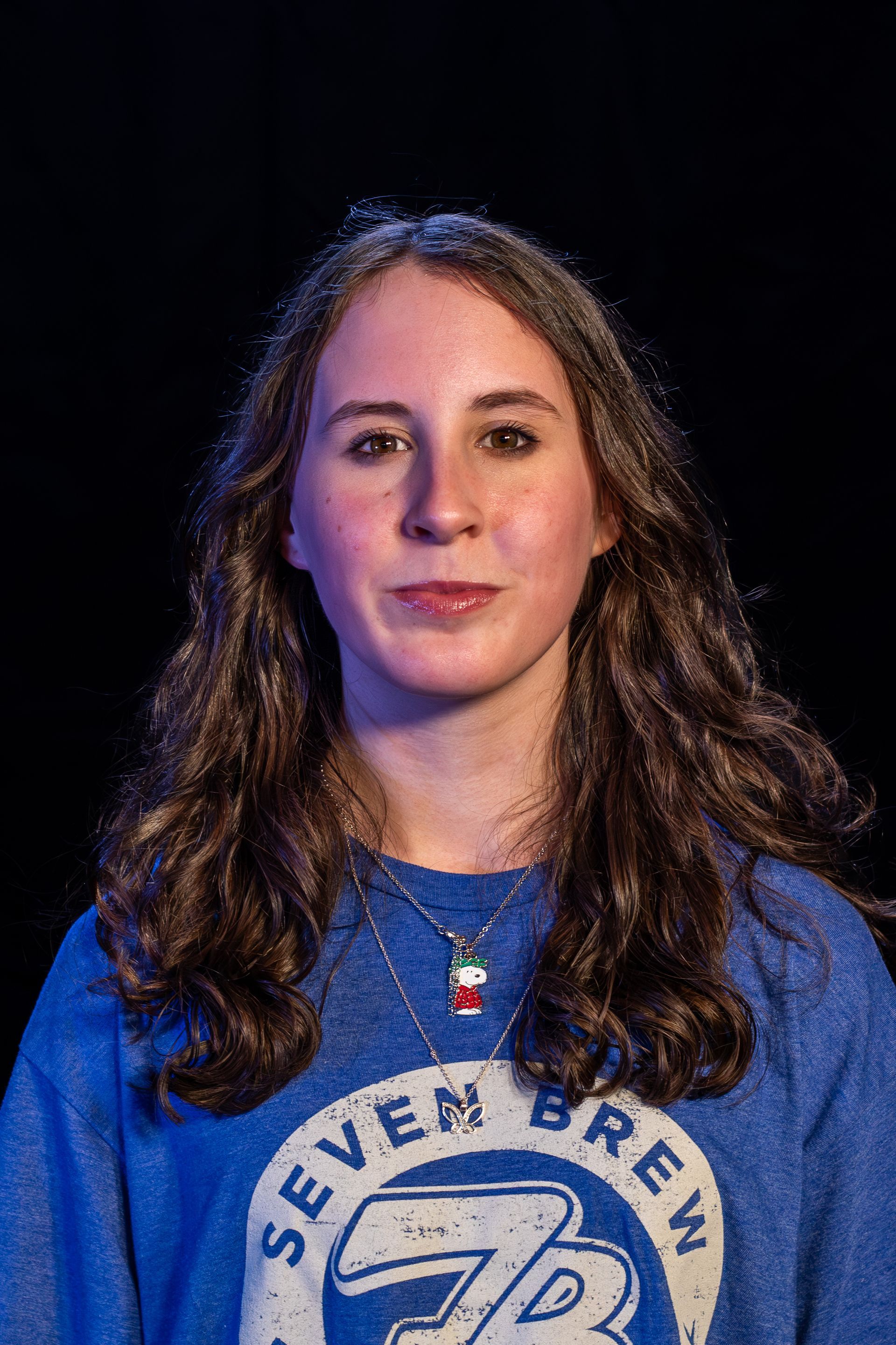 Woman with curly brown hair, wearing a blue sweatshirt and necklace, against a black background.