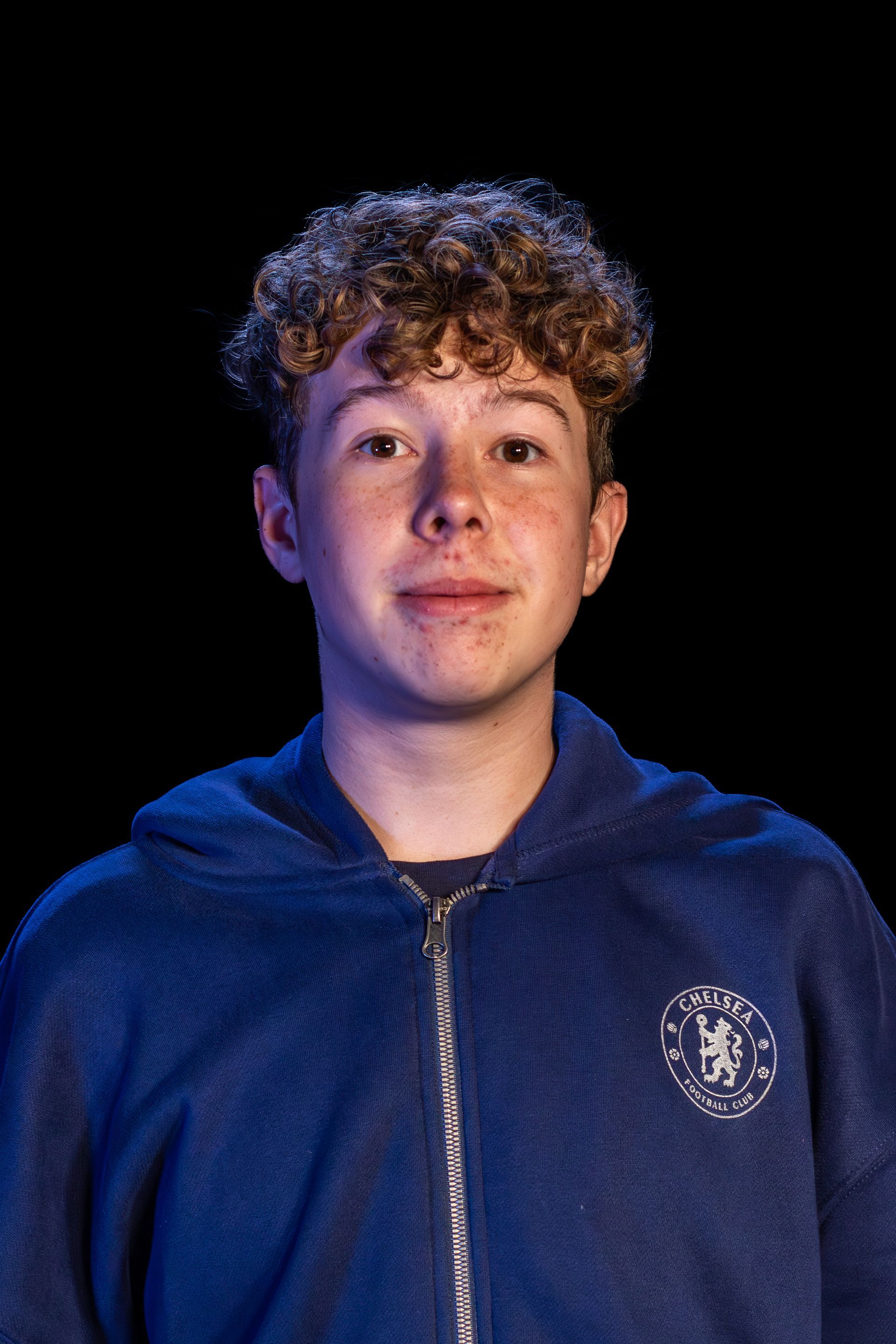 Teenager with curly hair, wearing a blue zip-up hoodie, smiling against a black background.