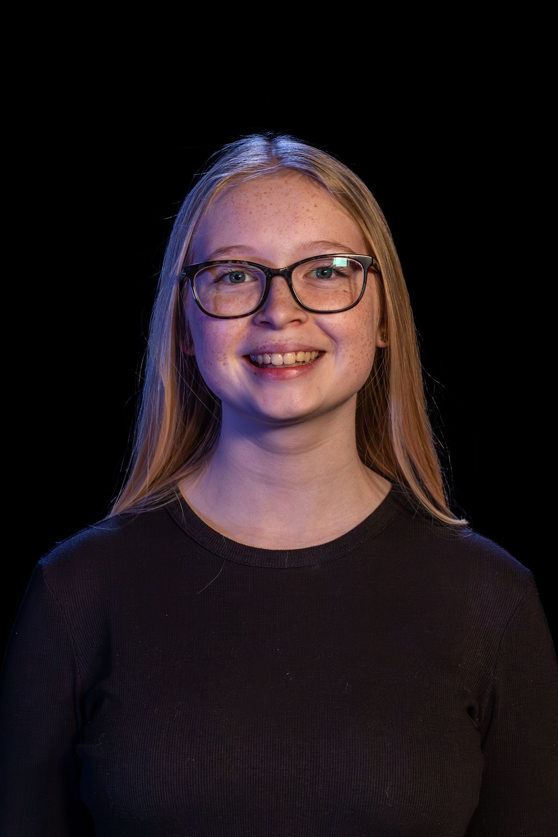 Smiling person with glasses, long blonde hair, and black shirt, lit with blue and purple light against a black backdrop.