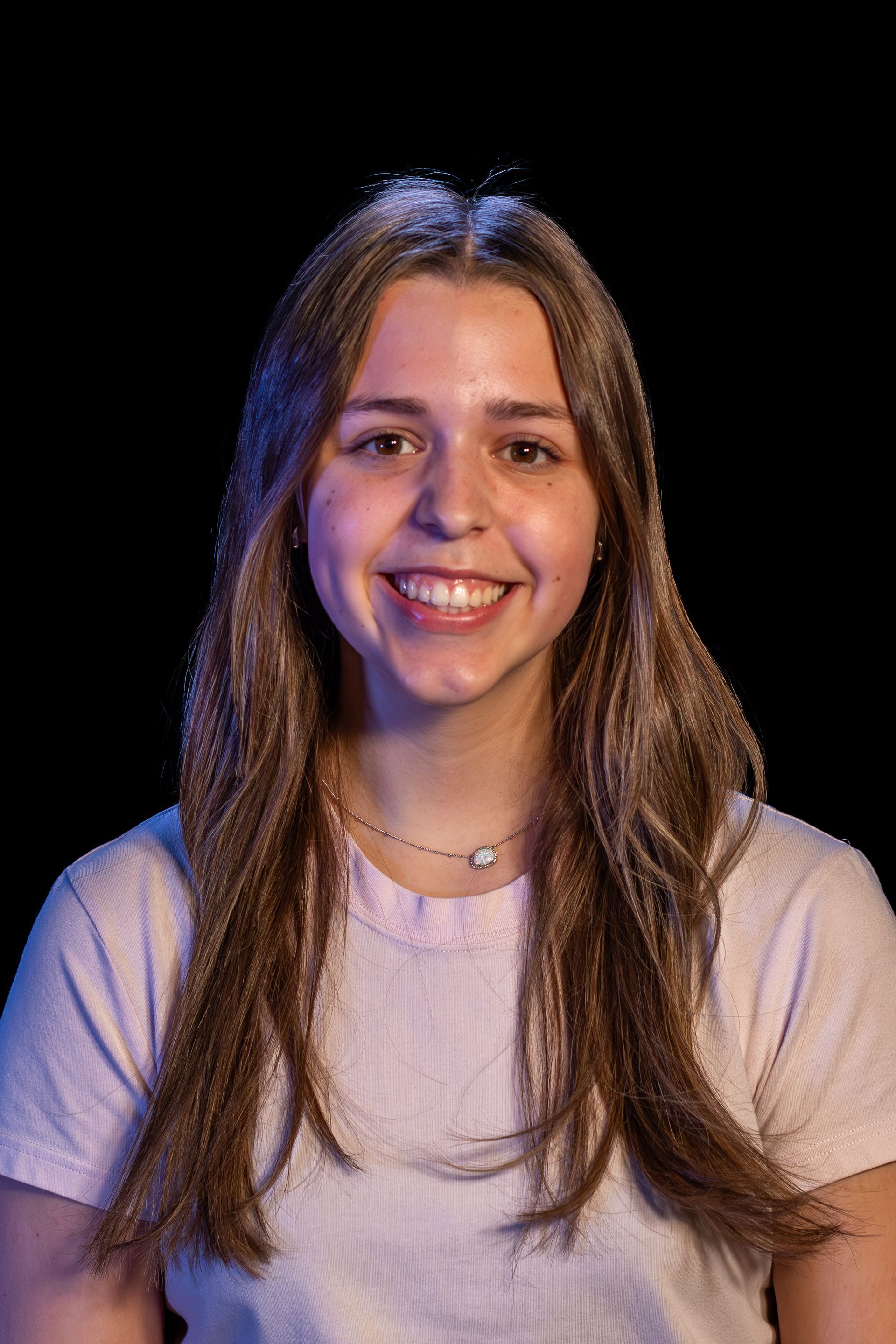 Smiling woman with long brown hair, wearing a light pink t-shirt, and a necklace. Black background.