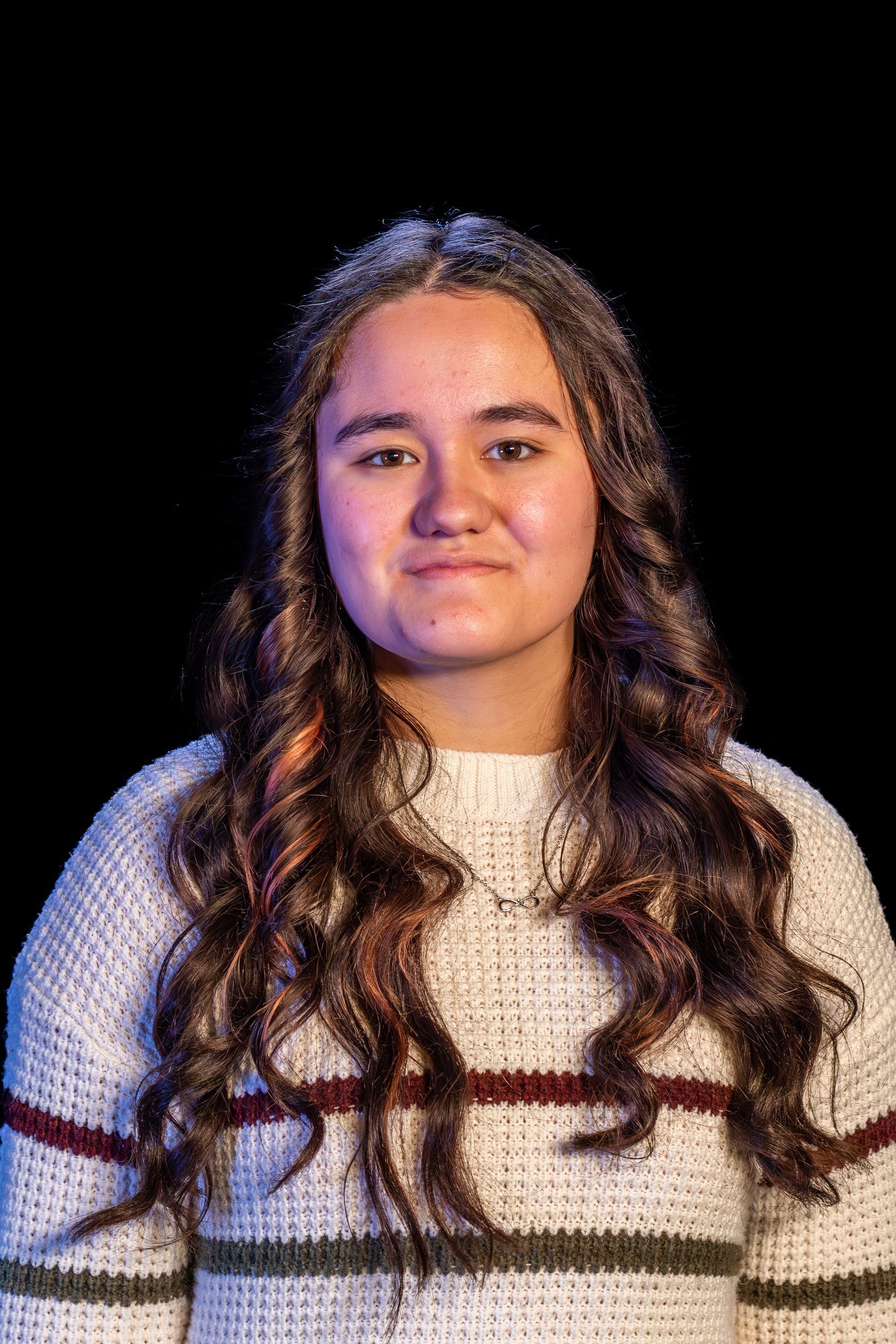Woman with long, wavy brown hair wearing a white sweater with maroon and green stripes, against a black backdrop.