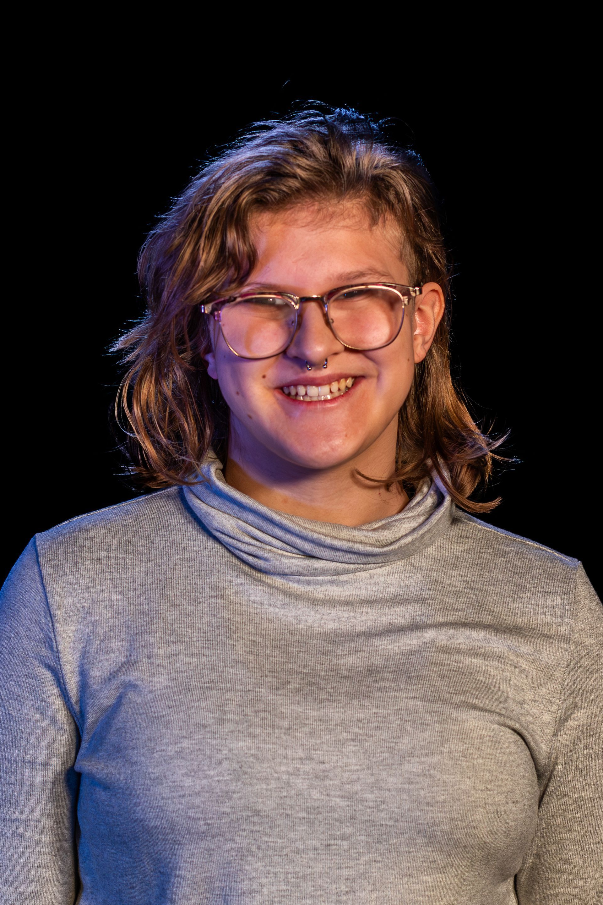 Woman with glasses smiling, wearing a gray turtleneck, against a black background.