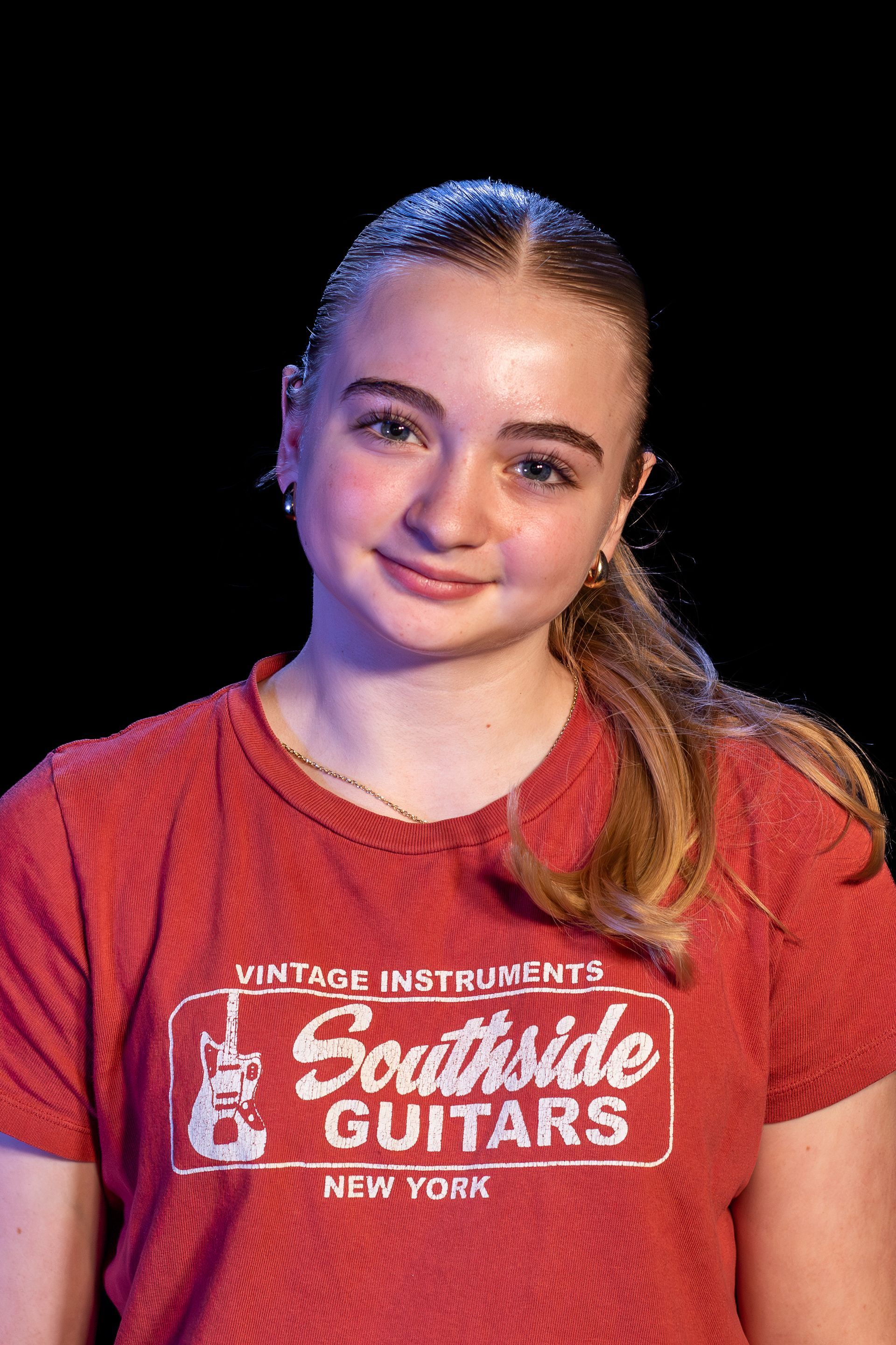Woman with light skin wearing a red t-shirt, smiling at the camera, with a black background.