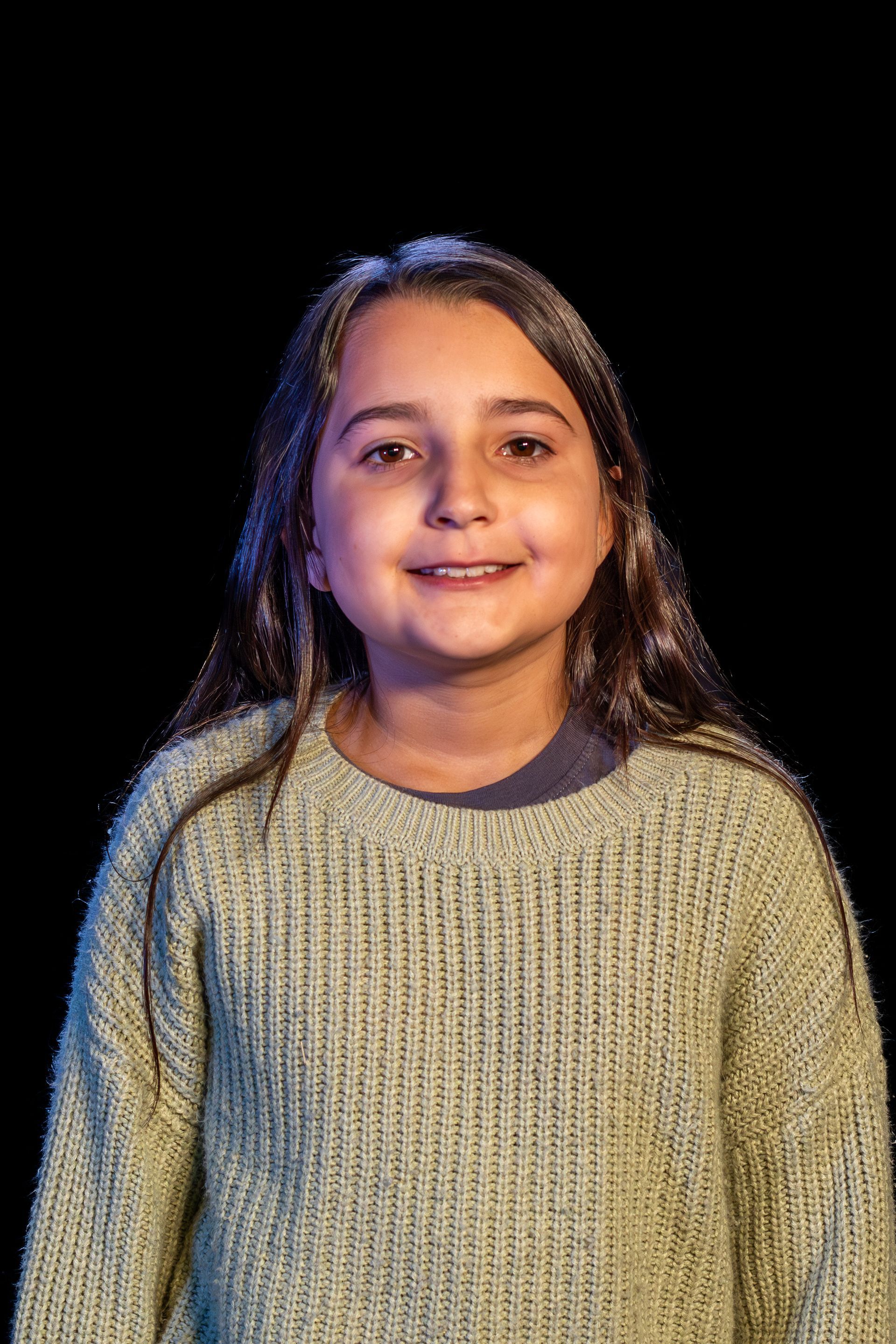 Young person with long hair, wearing a green sweater, smiling, against a black background.