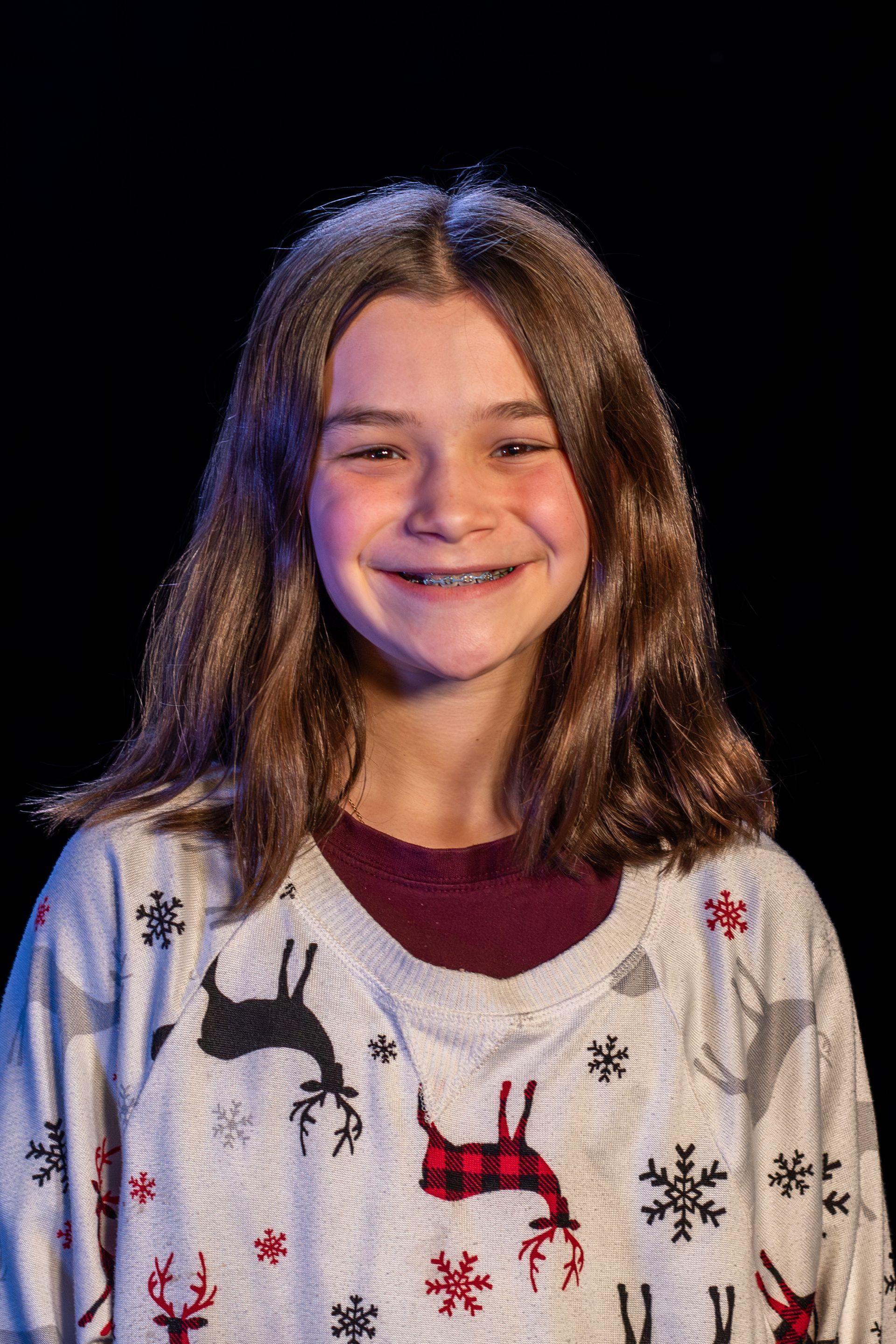 Girl smiling with braces, wearing a reindeer sweater, dark background.