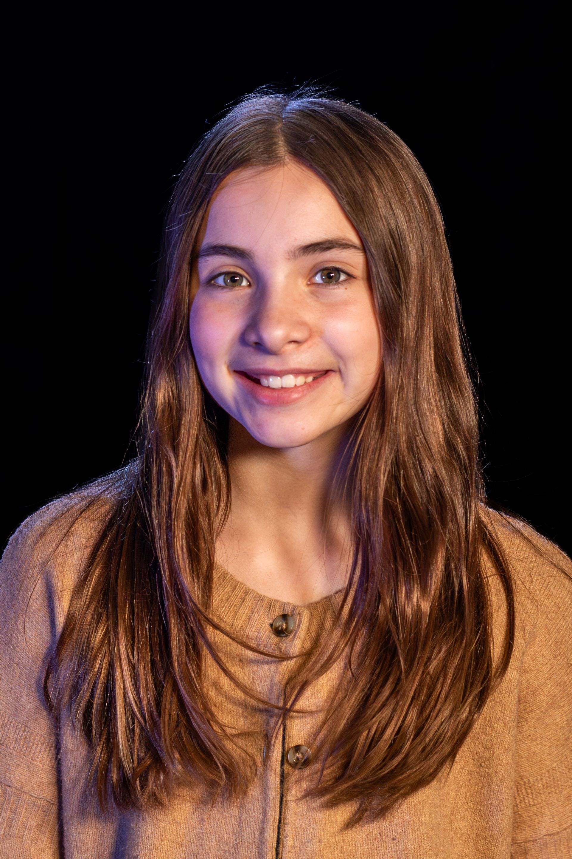 Girl with long brown hair smiles, wearing a tan sweater against a black background.