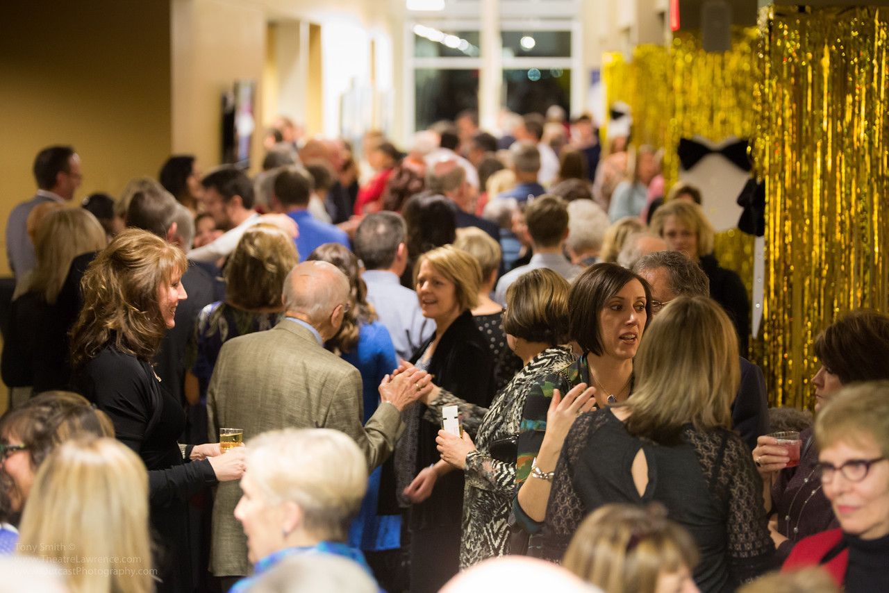 Crowd of people mingling at an event, some in formal attire. Gold decorations are visible in the background.
