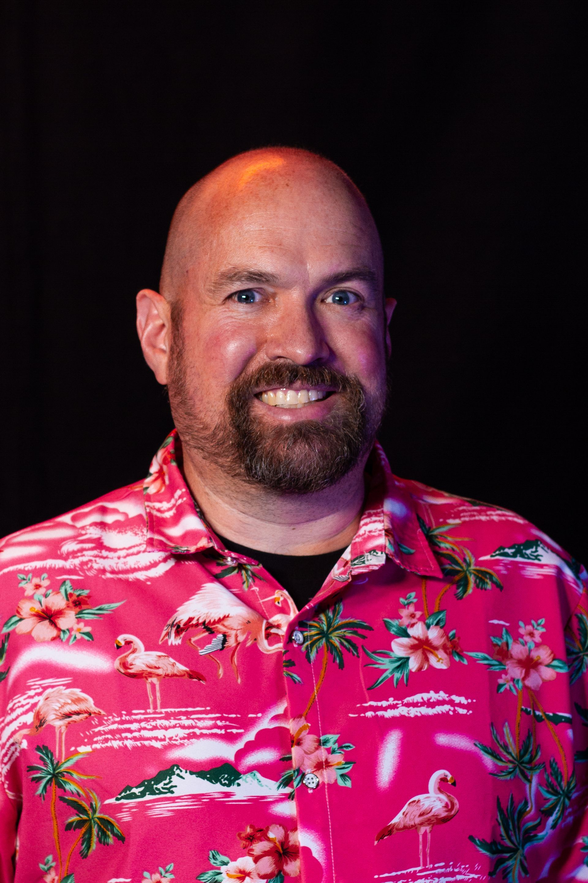 Man with beard smiling, wearing a pink floral shirt with flamingos, against a dark background.