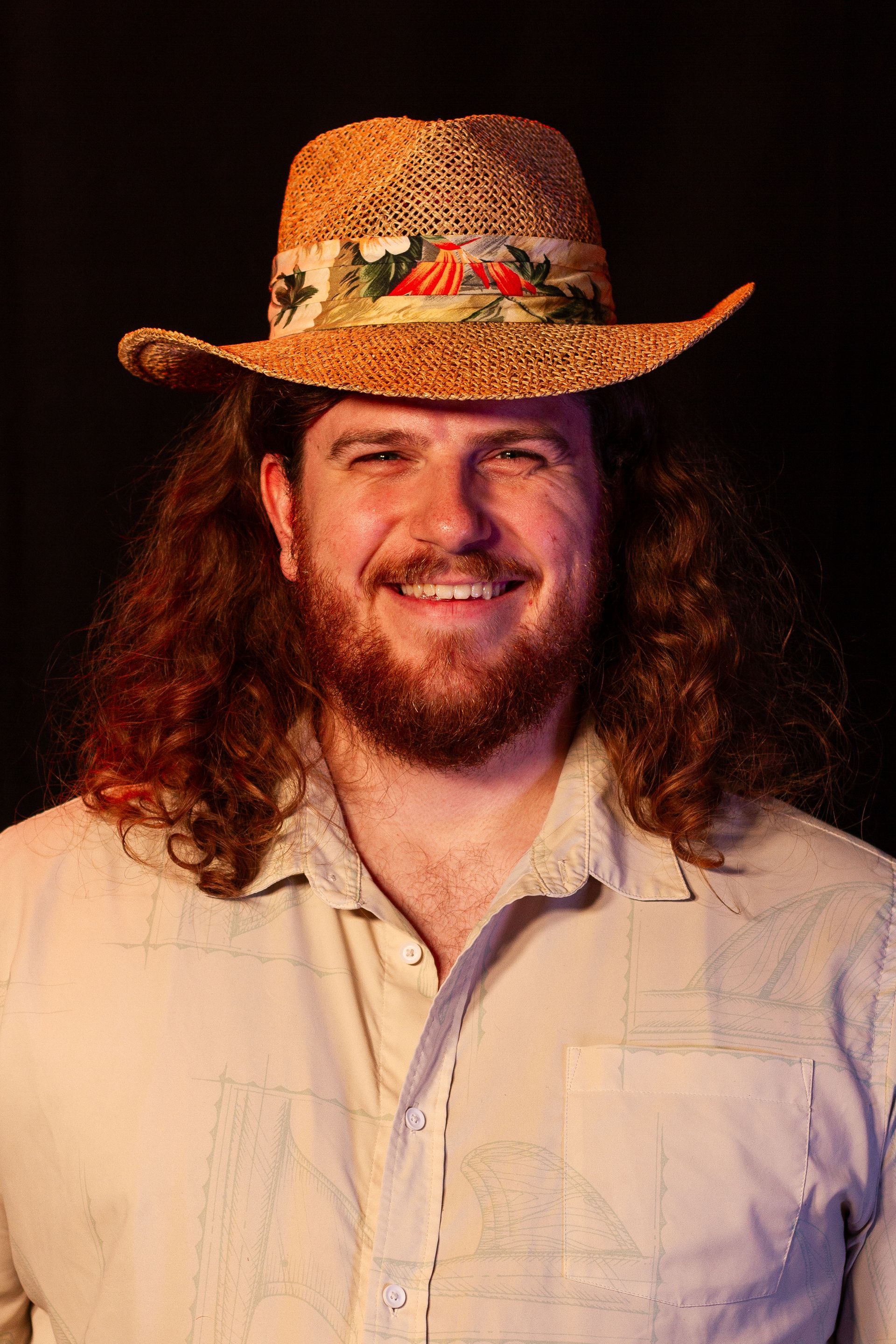 Man with long curly hair, beard, and straw hat smiling, wearing a light button-down shirt.