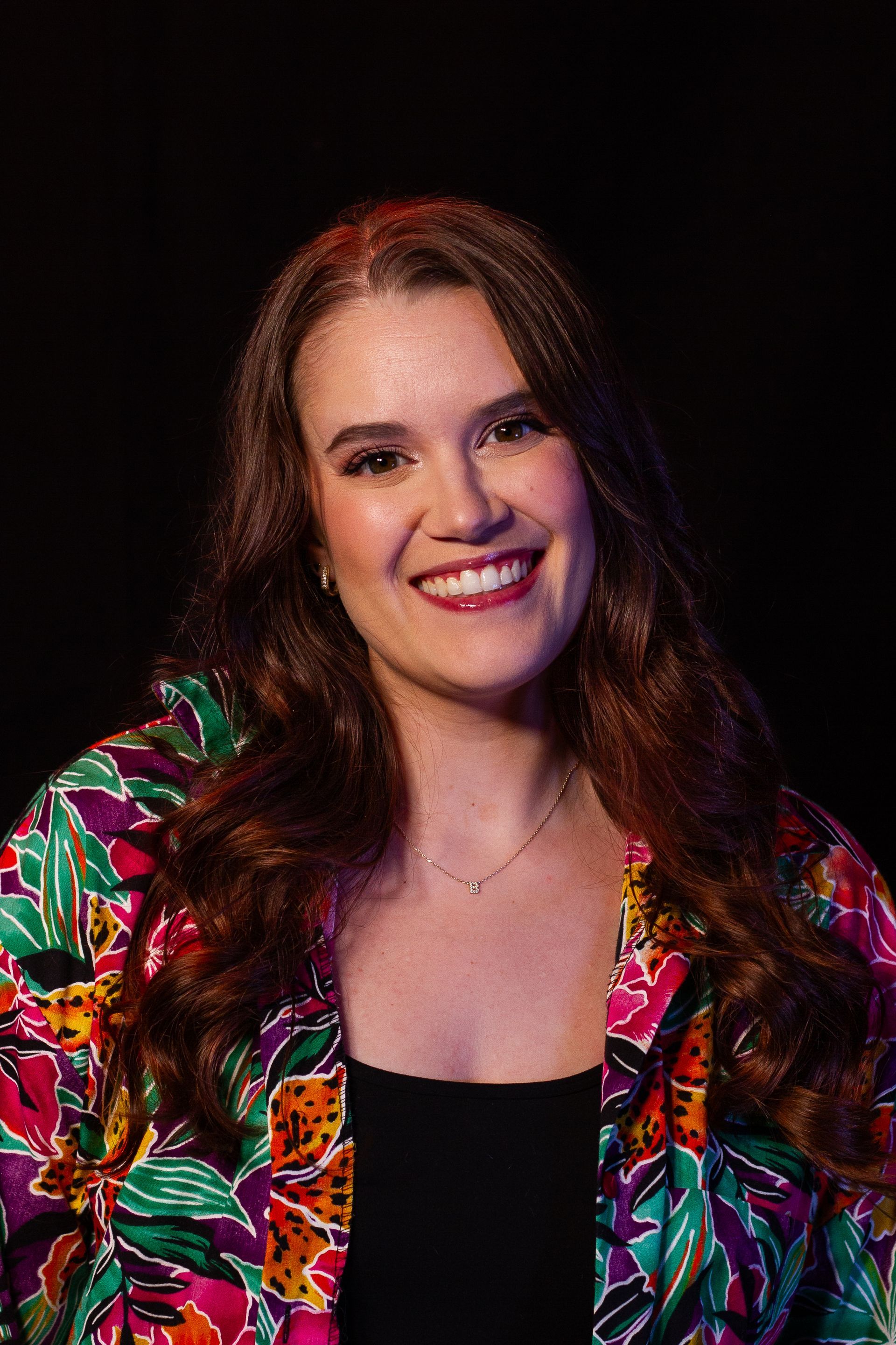 Smiling woman with long red hair, wearing a colorful floral jacket, against a black background.