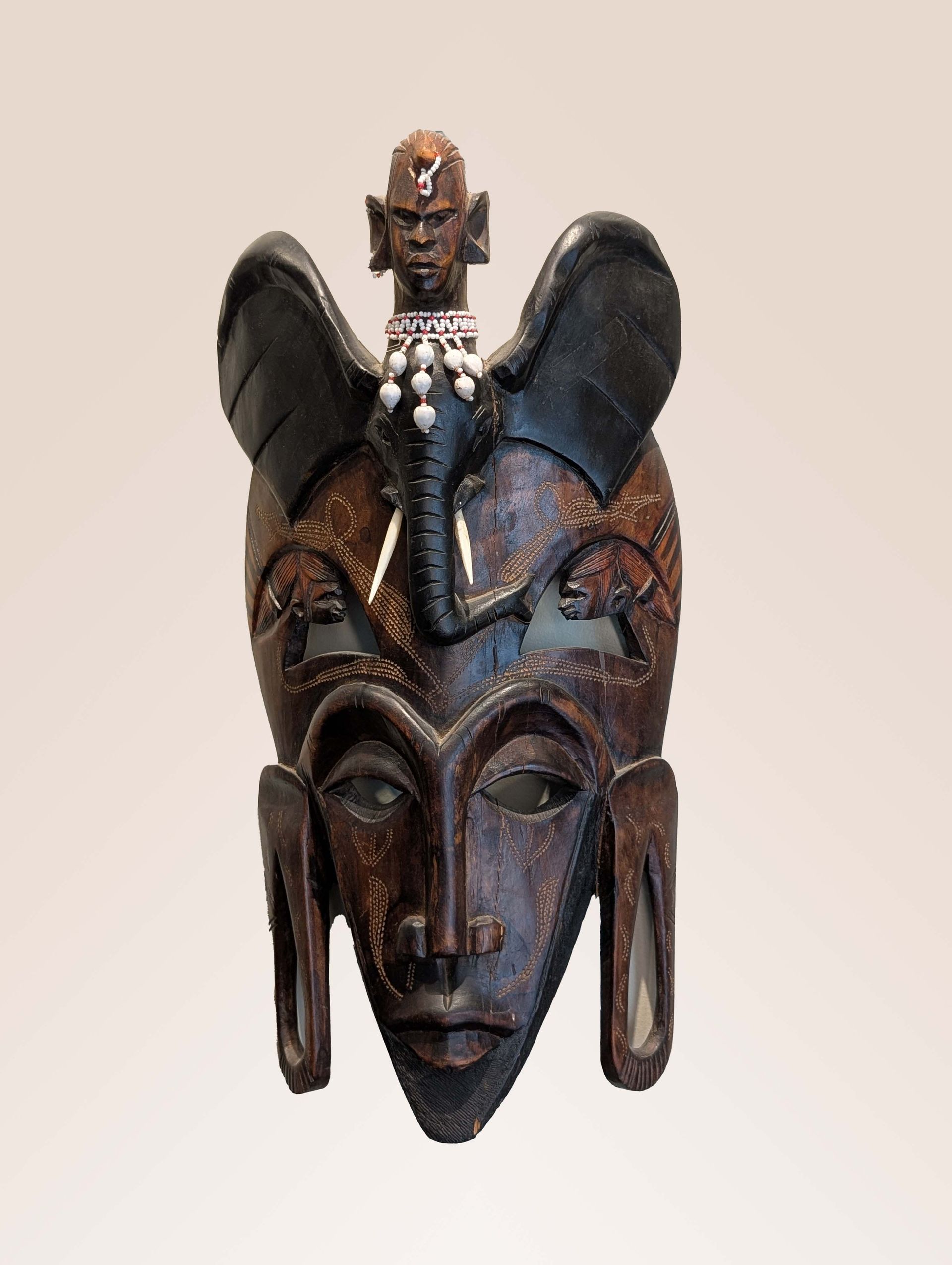 Wooden African mask with elephant ears and trunk, and human-like facial features, with a small human figure on top.