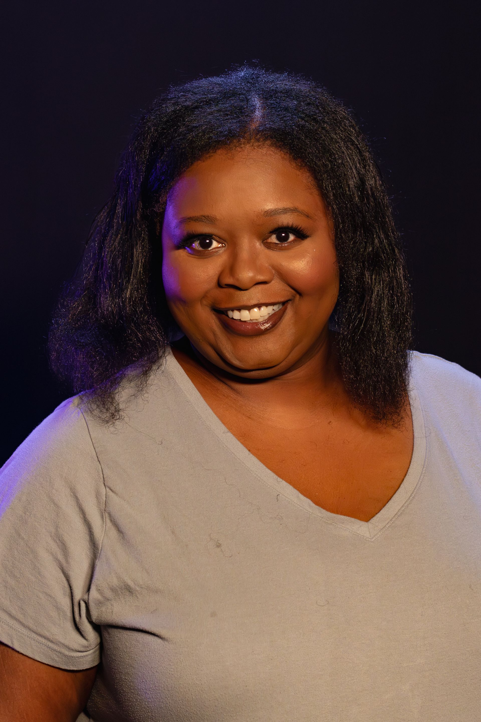 Woman with dark hair smiles, wearing a grey v-neck shirt, lit by purple and blue light.