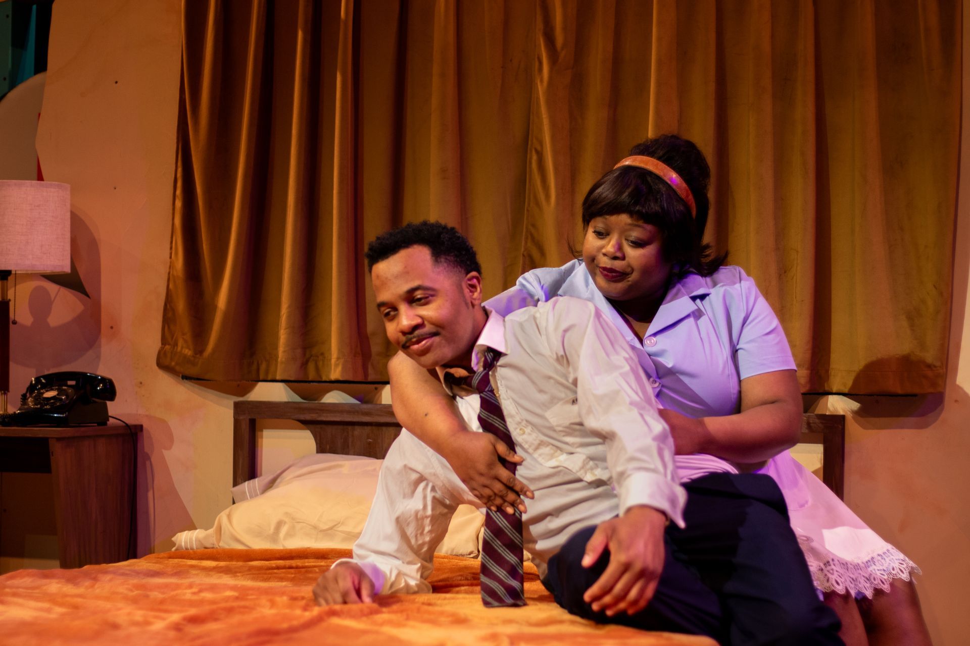 Two people in a hotel room. One leans on the bed, the other hugs their shoulder. Beige and brown tones.