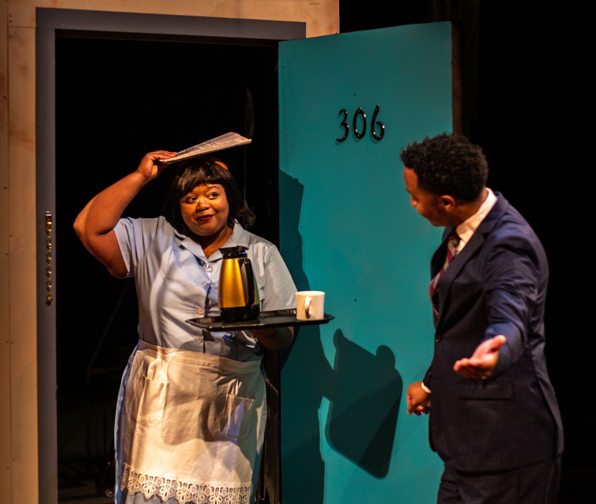 A person in a waitress uniform holding a tray greets a person in a suit by a hotel room door.