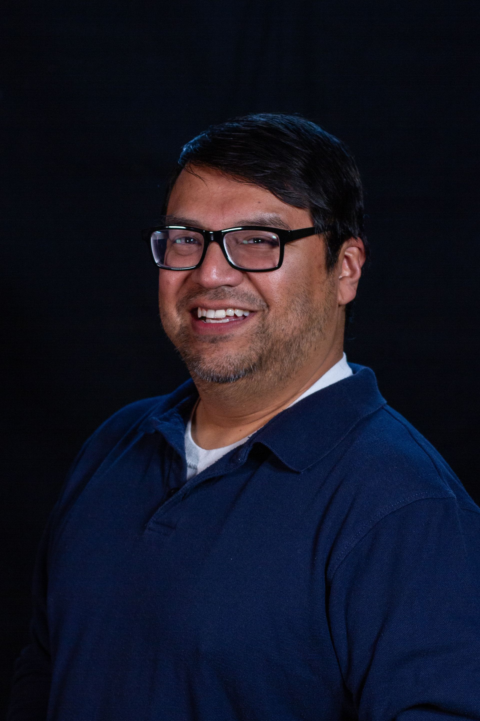 Man in glasses, blue shirt, smiling, against a dark background.
