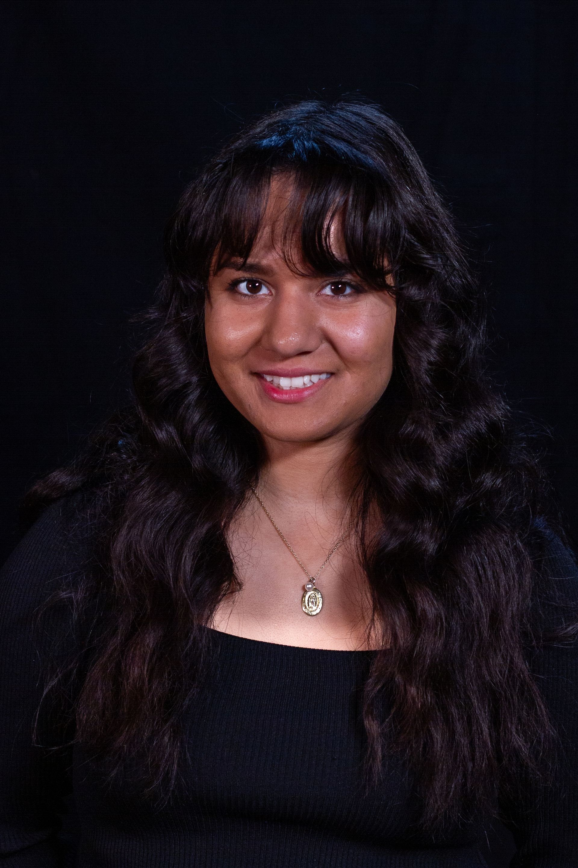 Woman with long, wavy dark hair smiling, wearing a black top and necklace, against a black background.