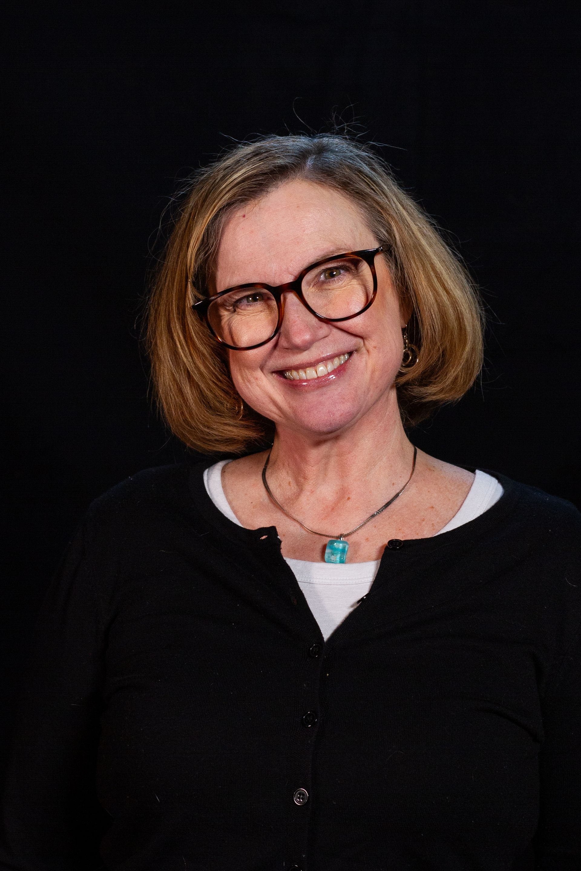 Woman with glasses, smiling, wearing a black cardigan over a white shirt, against a black background.