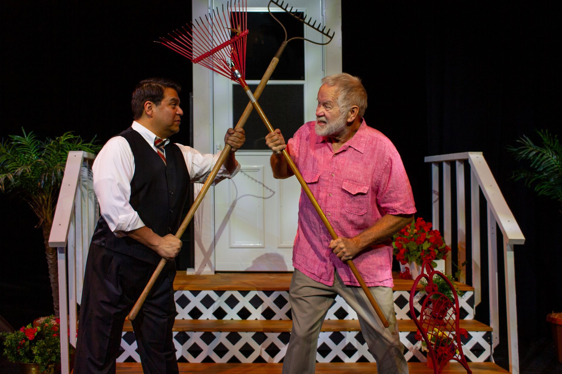 Two men, one in a vest, one in pink shirt, fight with garden rakes on a porch.