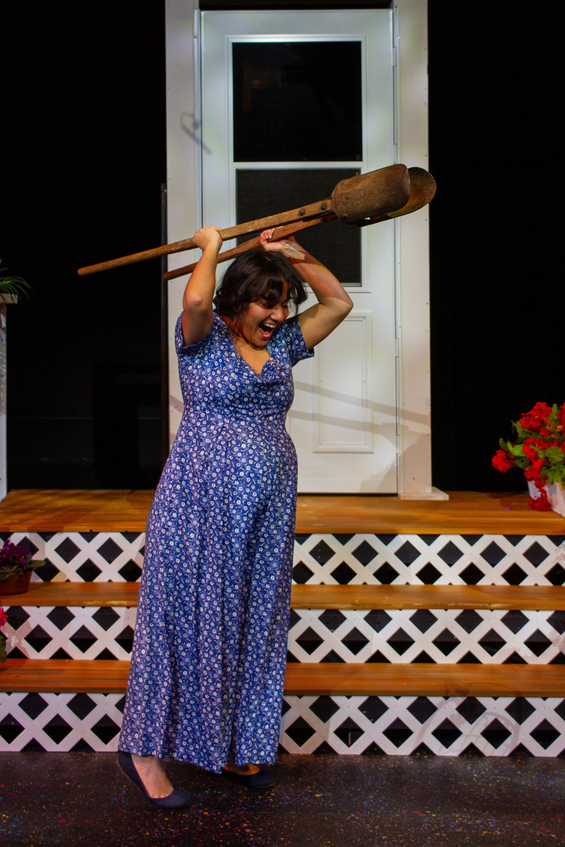 Woman in blue dress holds a large wooden paddle overhead, grinning on a porch.