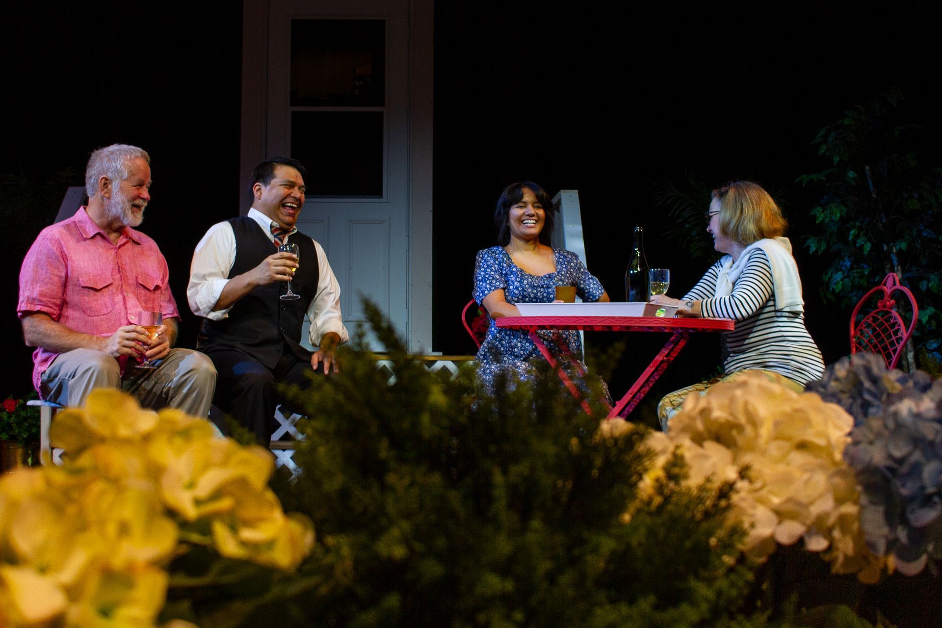 Four people laughing on a patio, near a red table and flowers. Daytime.