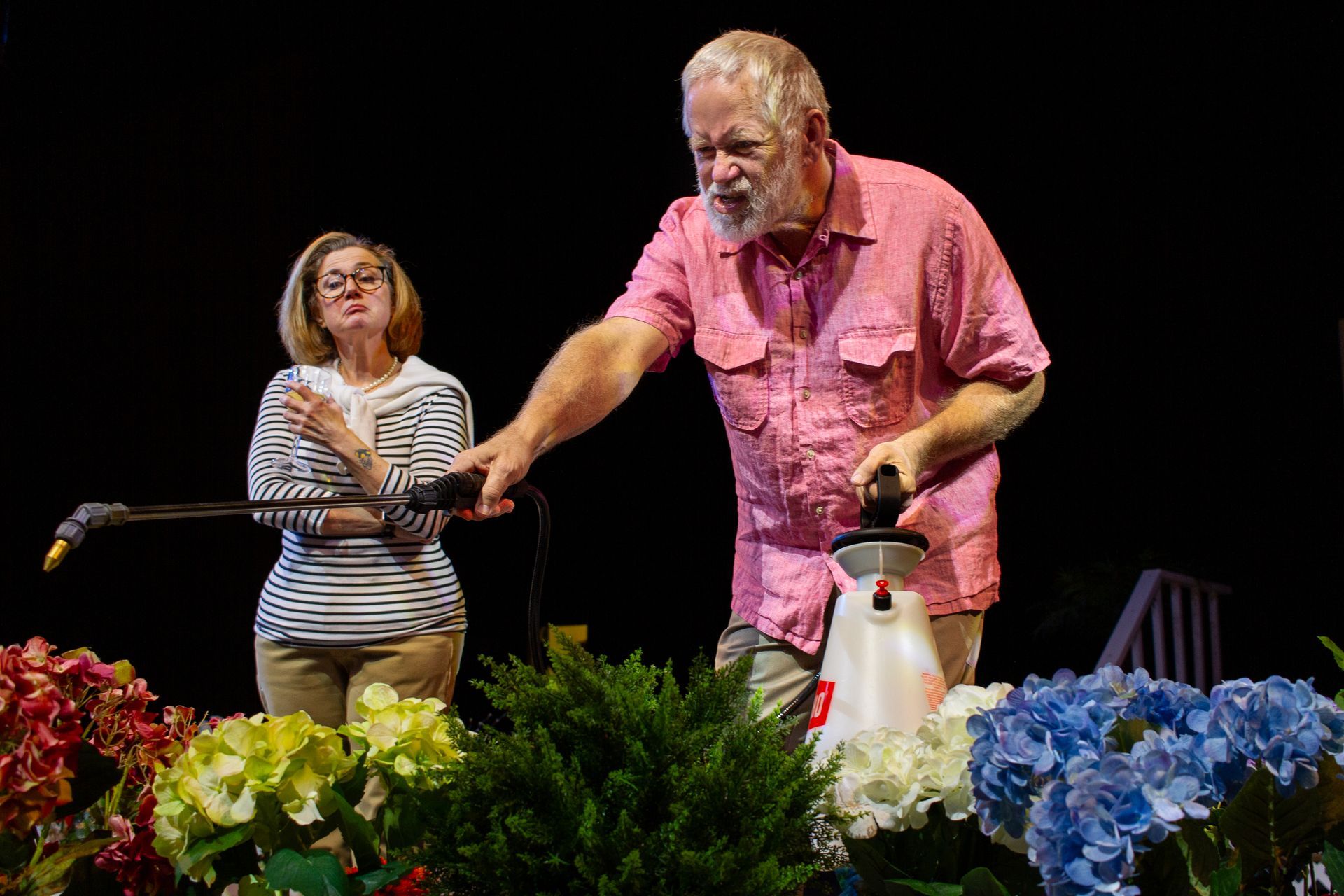 A man sprays plants as a woman watches, both outdoors.  Bright flowers surround them.