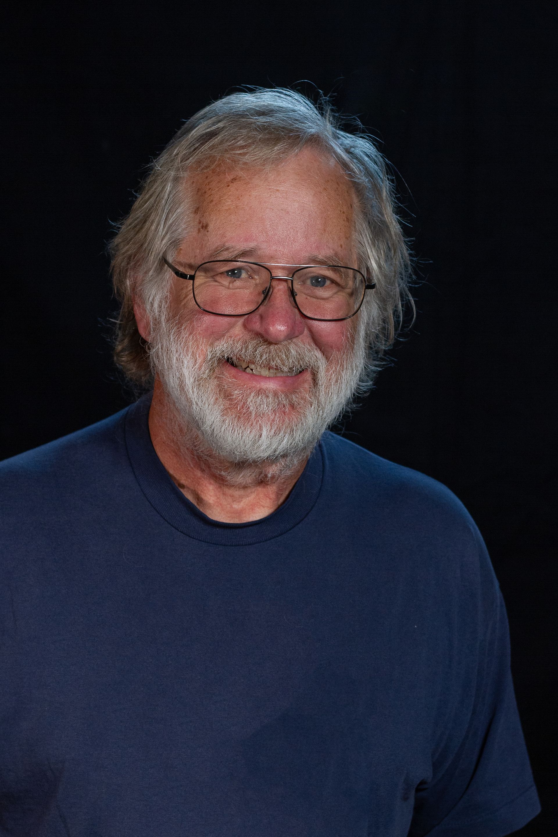 Older man with gray hair and beard wearing glasses and a blue shirt, smiling against a dark background.