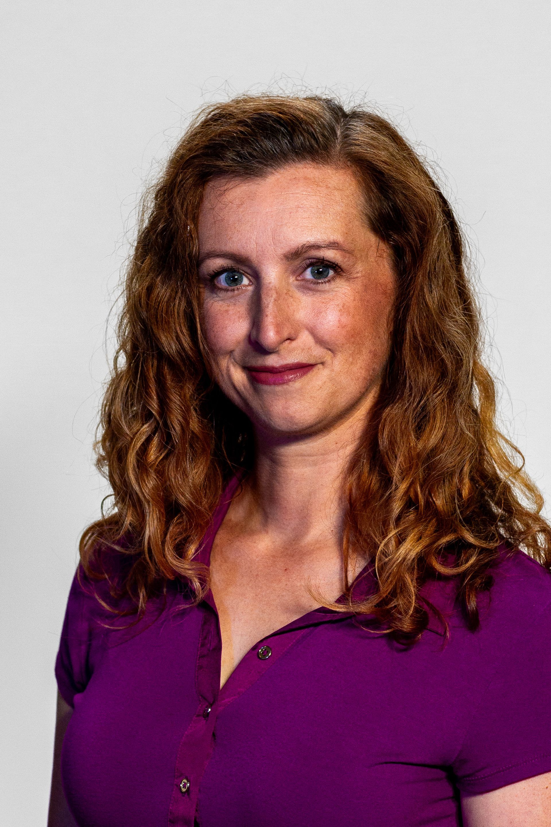 Woman with curly red hair, wearing a purple shirt, smiling, against a white background.