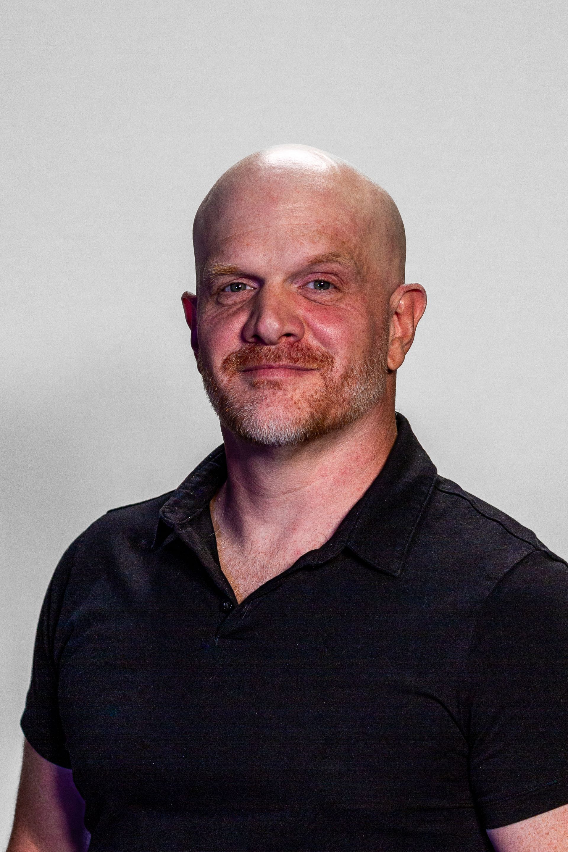 Bald man with red beard, smiling, wearing a black polo shirt, against a white background.