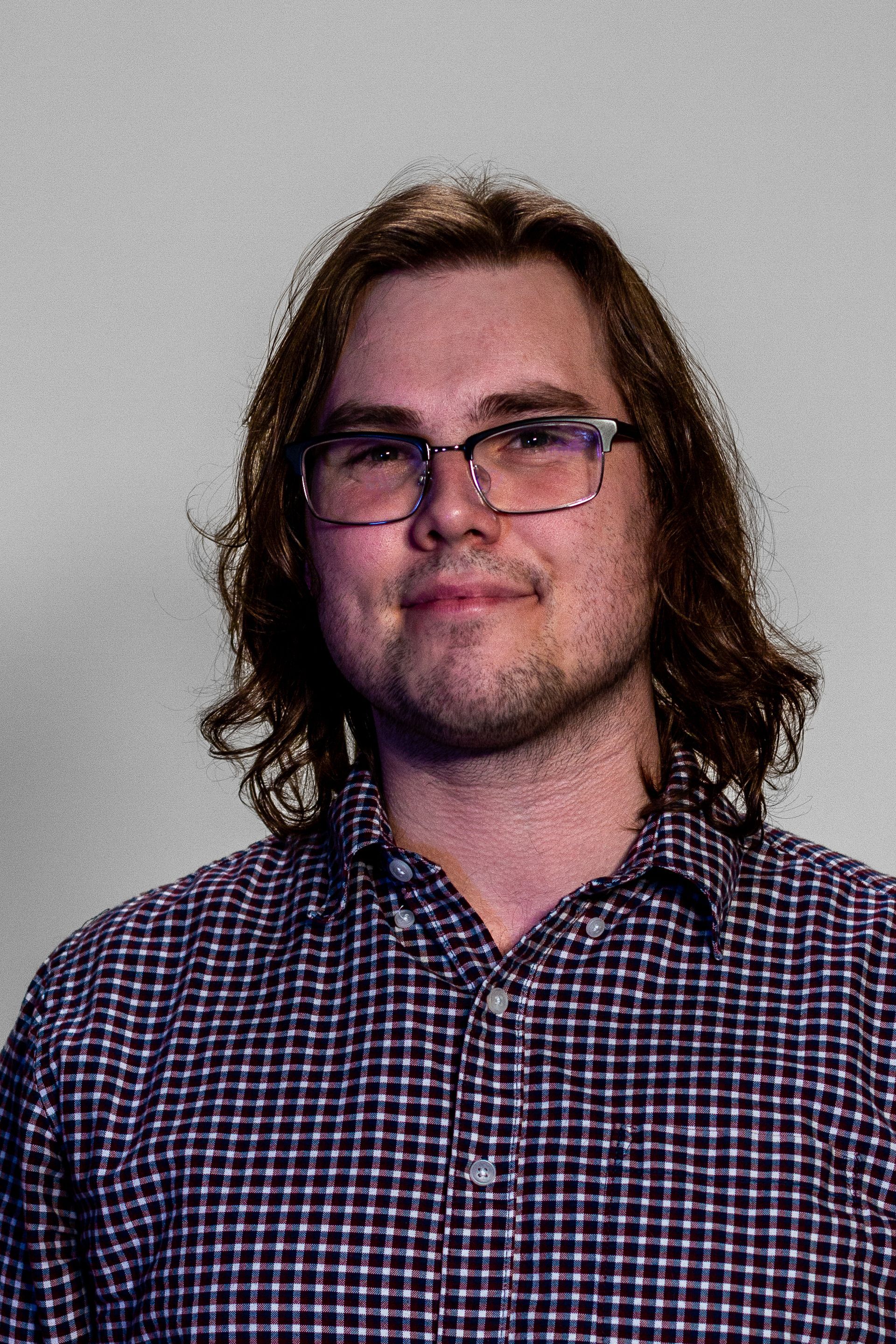 Man with long hair and glasses, wearing a checkered shirt, smiling, standing against a gray backdrop.