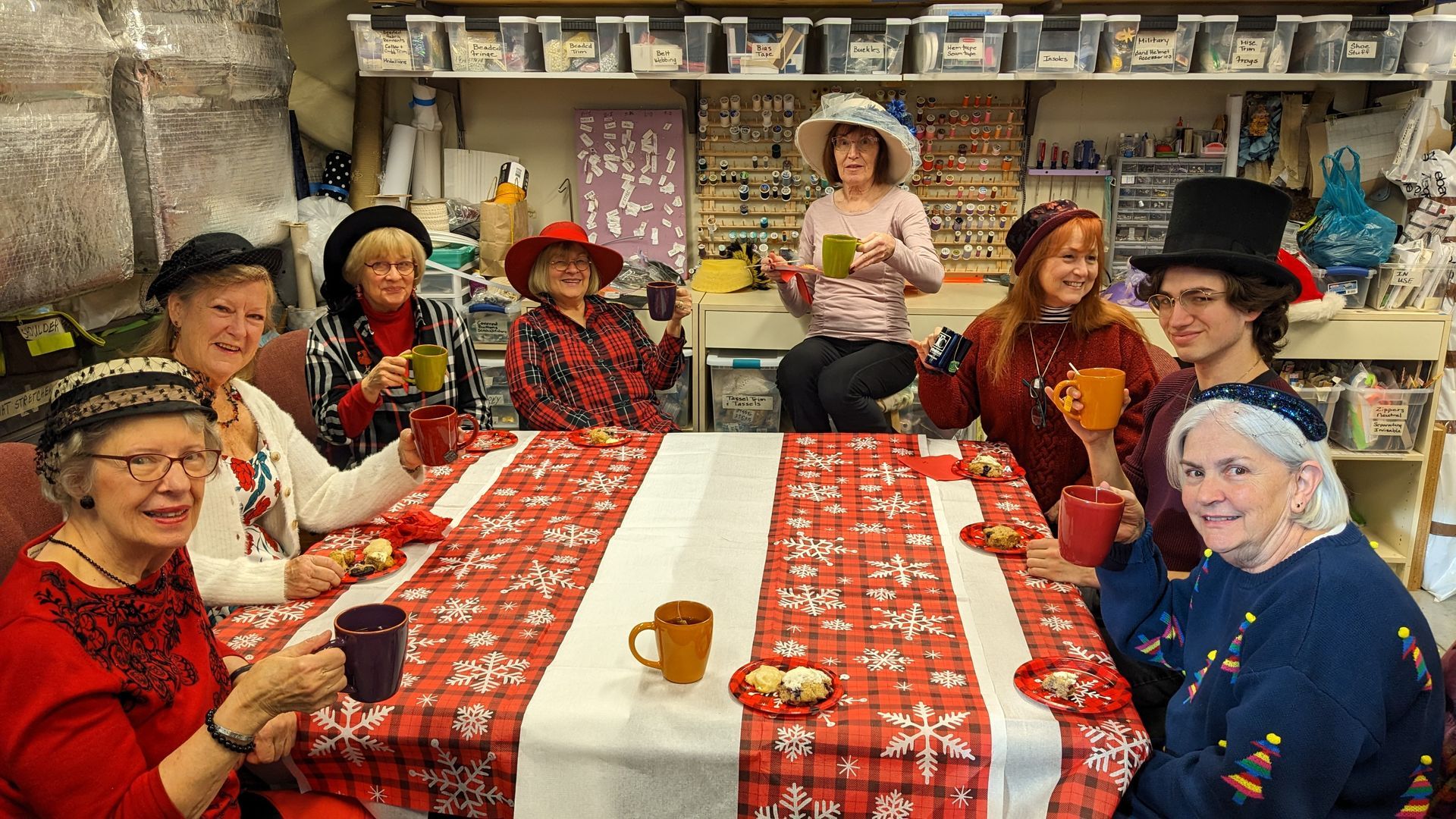 Group of people in hats at a table, toasting with mugs. Festive red & white tablecloth, cheerful expressions.