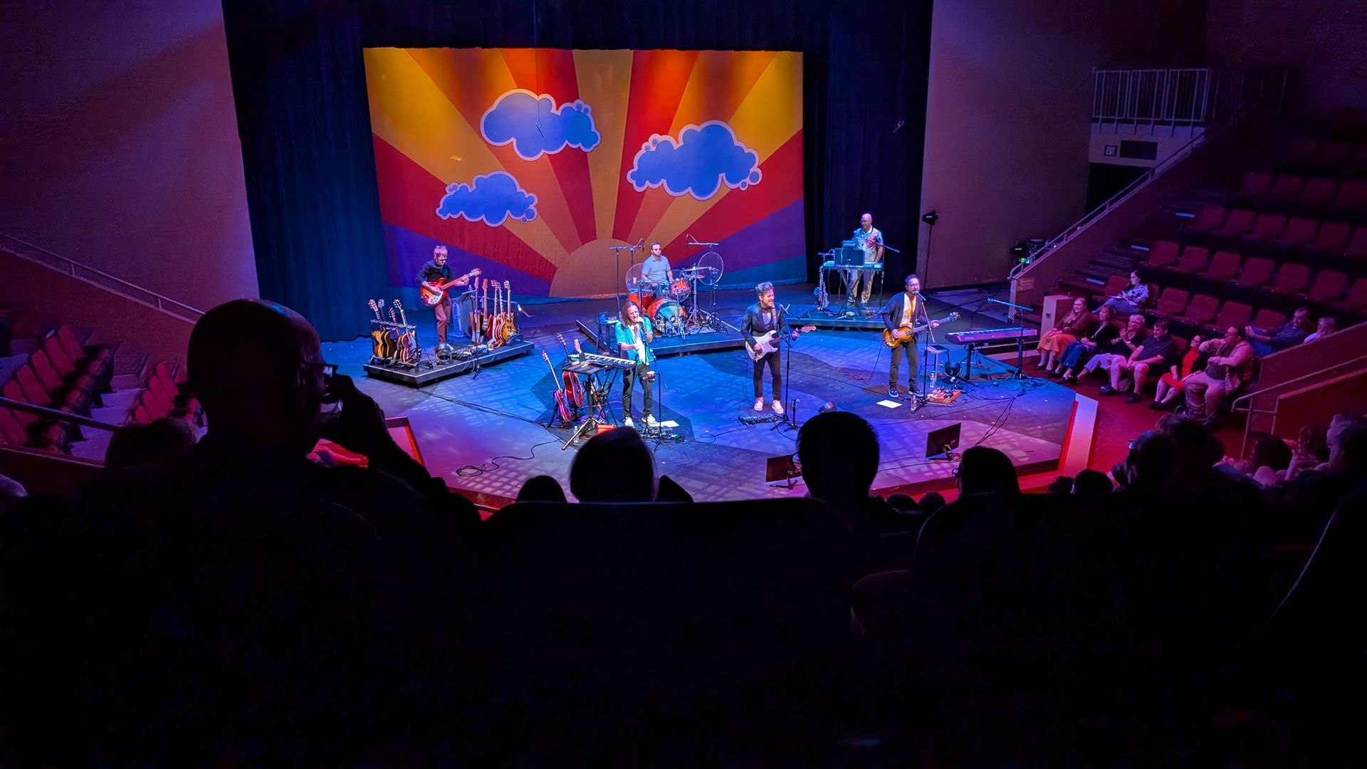 A band performs on stage with a colorful backdrop depicting a sunrise and clouds, viewed from the audience.