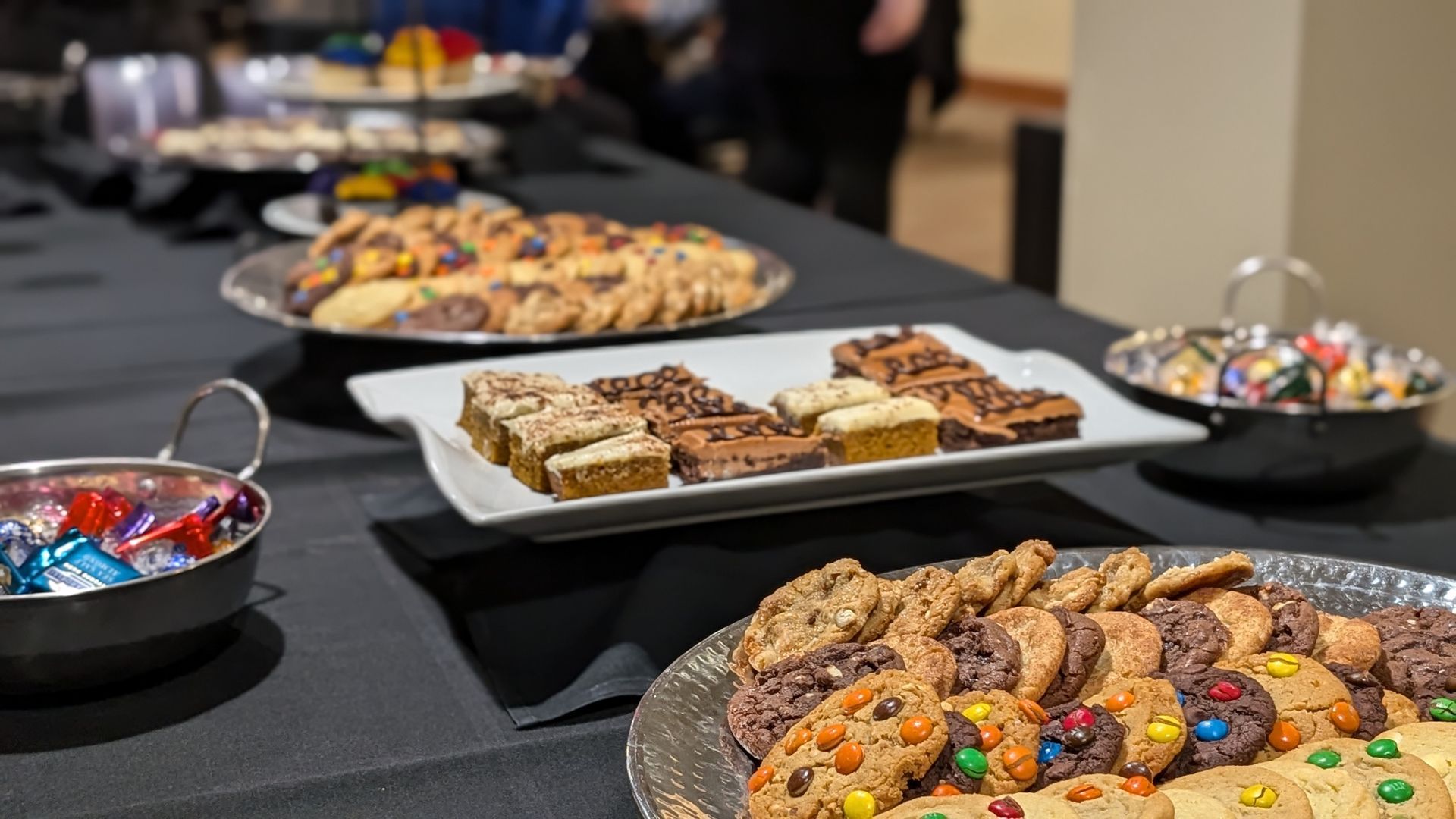 Table with desserts: cookies, brownies, and candies. Black tablecloth. Indoor setting.