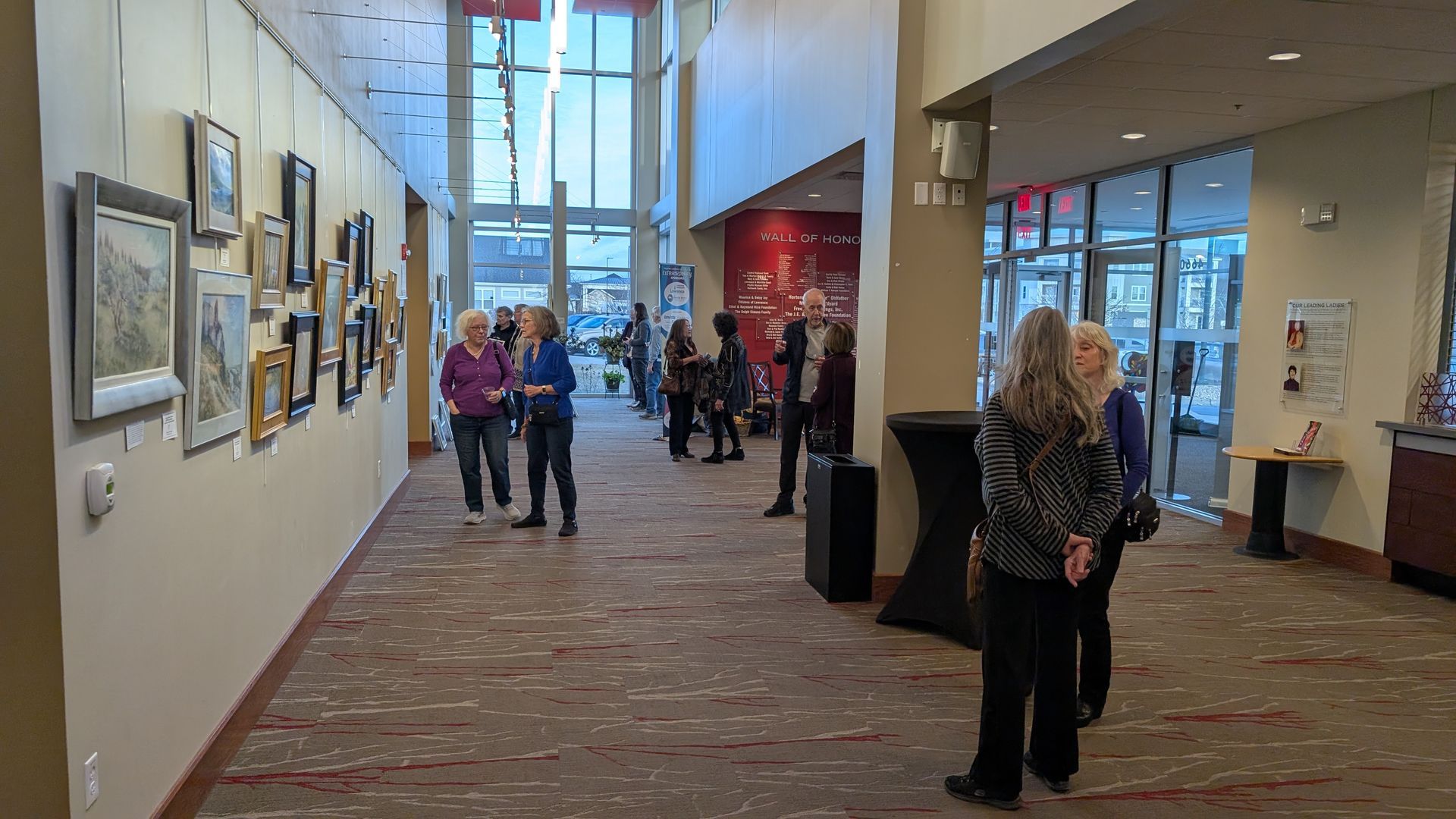 People view art in a gallery. Paintings line the wall. Natural light streams in from large windows.
