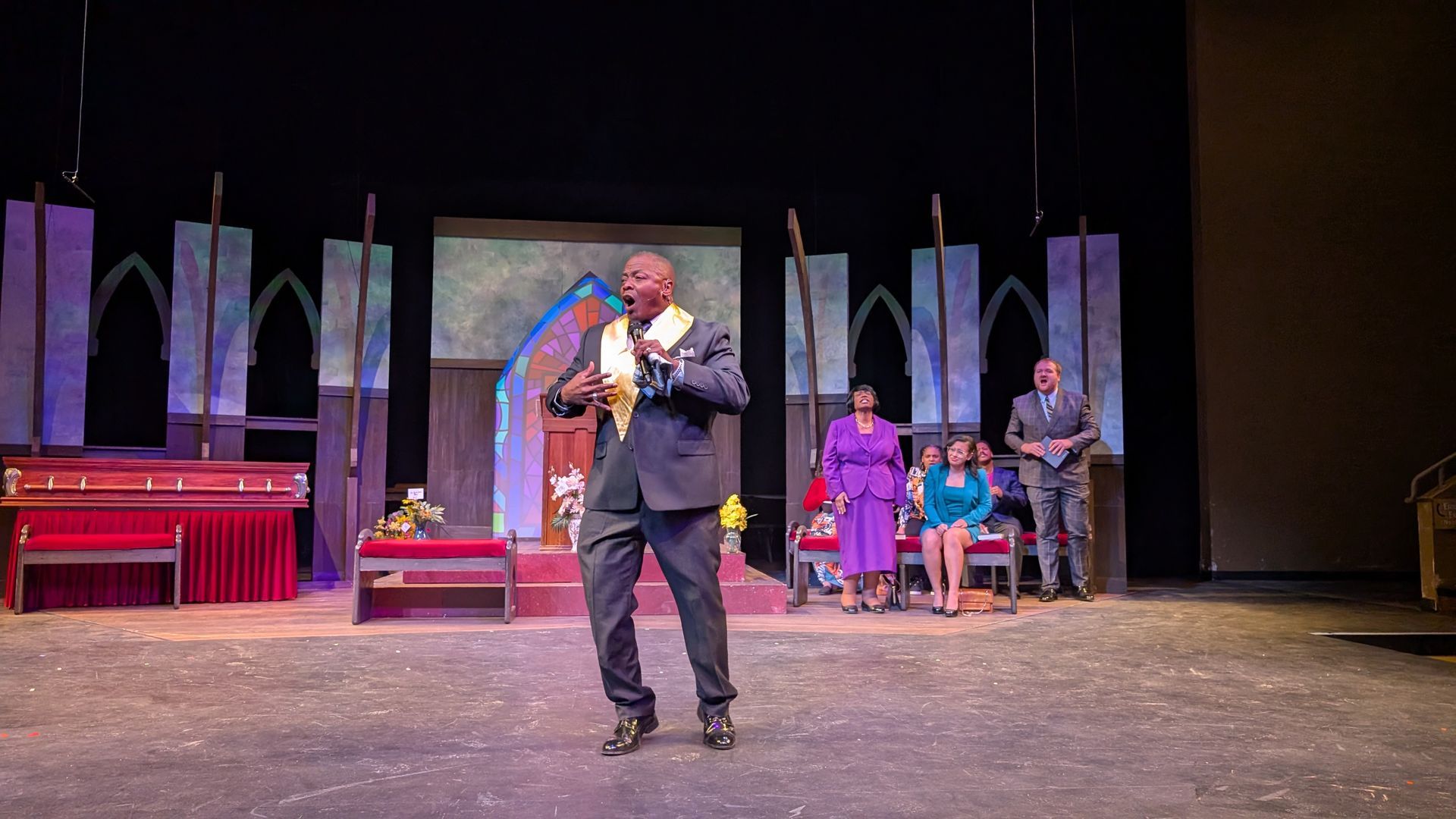 A man in a suit speaks on a stage decorated as a church, with other people in the background.