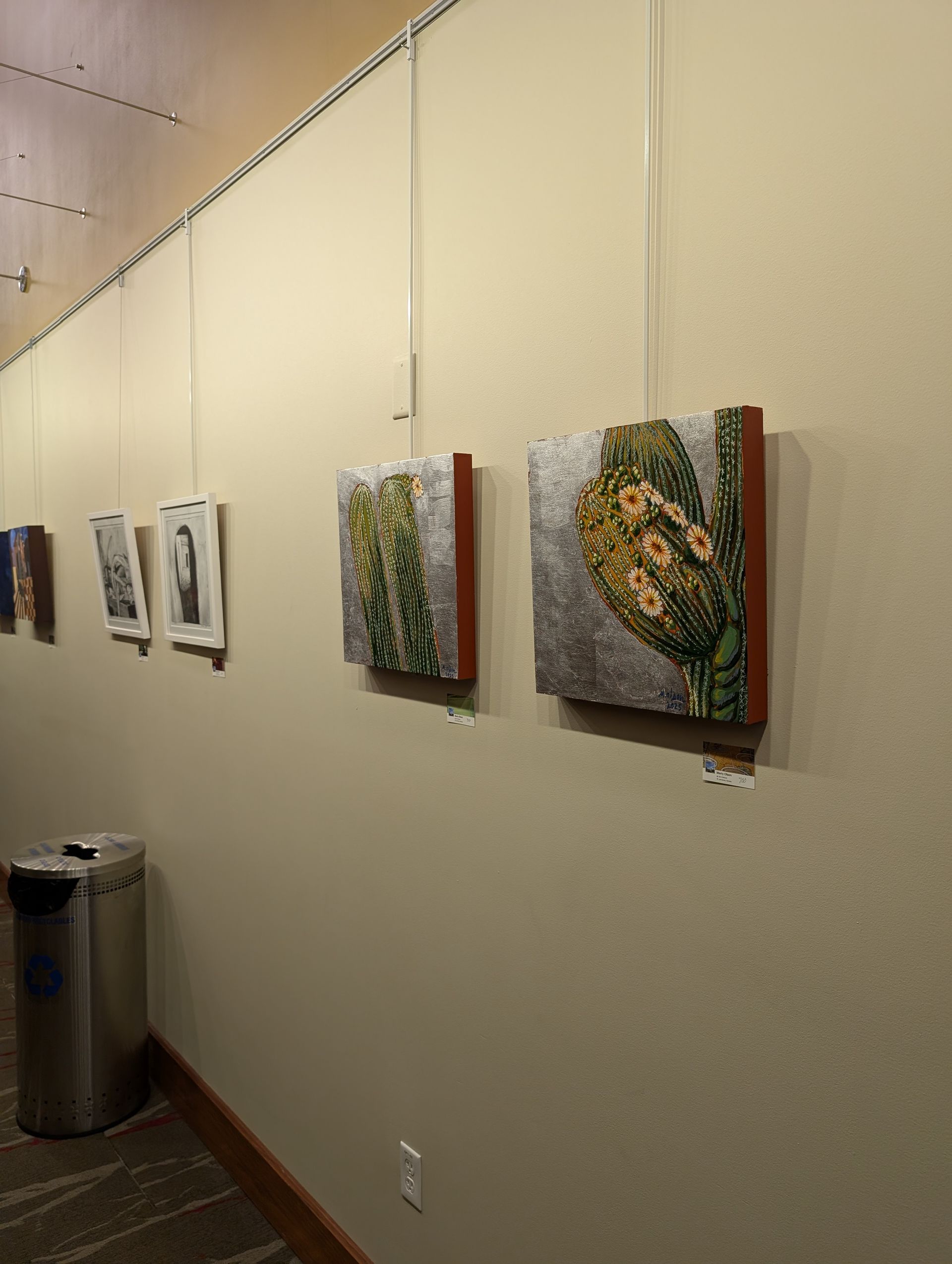 Artwork display in a gallery, with several framed pieces hanging on a beige wall.