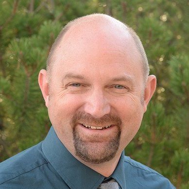 A bald man with a beard wearing a blue shirt and tie is smiling.