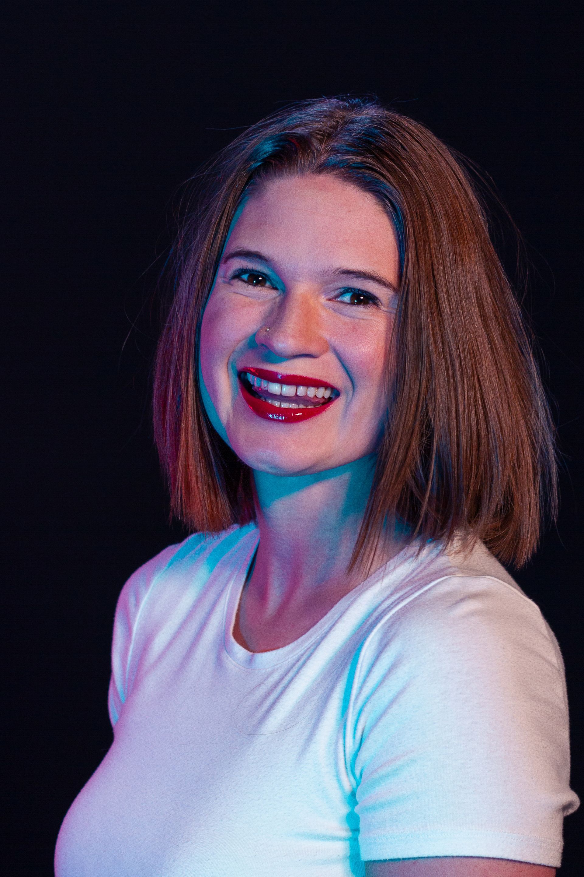 Woman with shoulder-length brown hair wearing a white shirt, smiling with red lipstick, lit with blue and pink light.