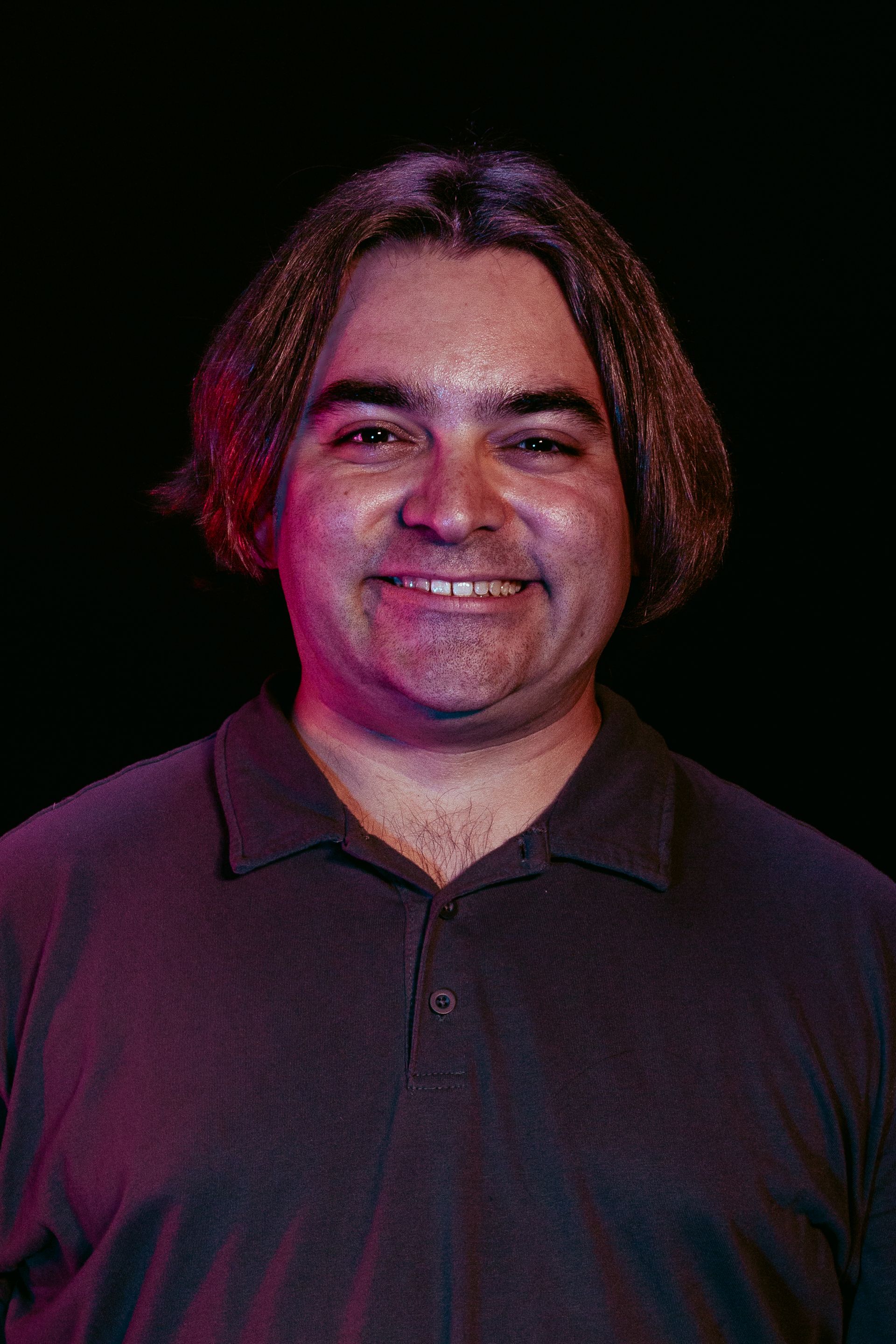 Man with brown hair and a smile, wearing a dark shirt, lit by red and blue light on a black background.