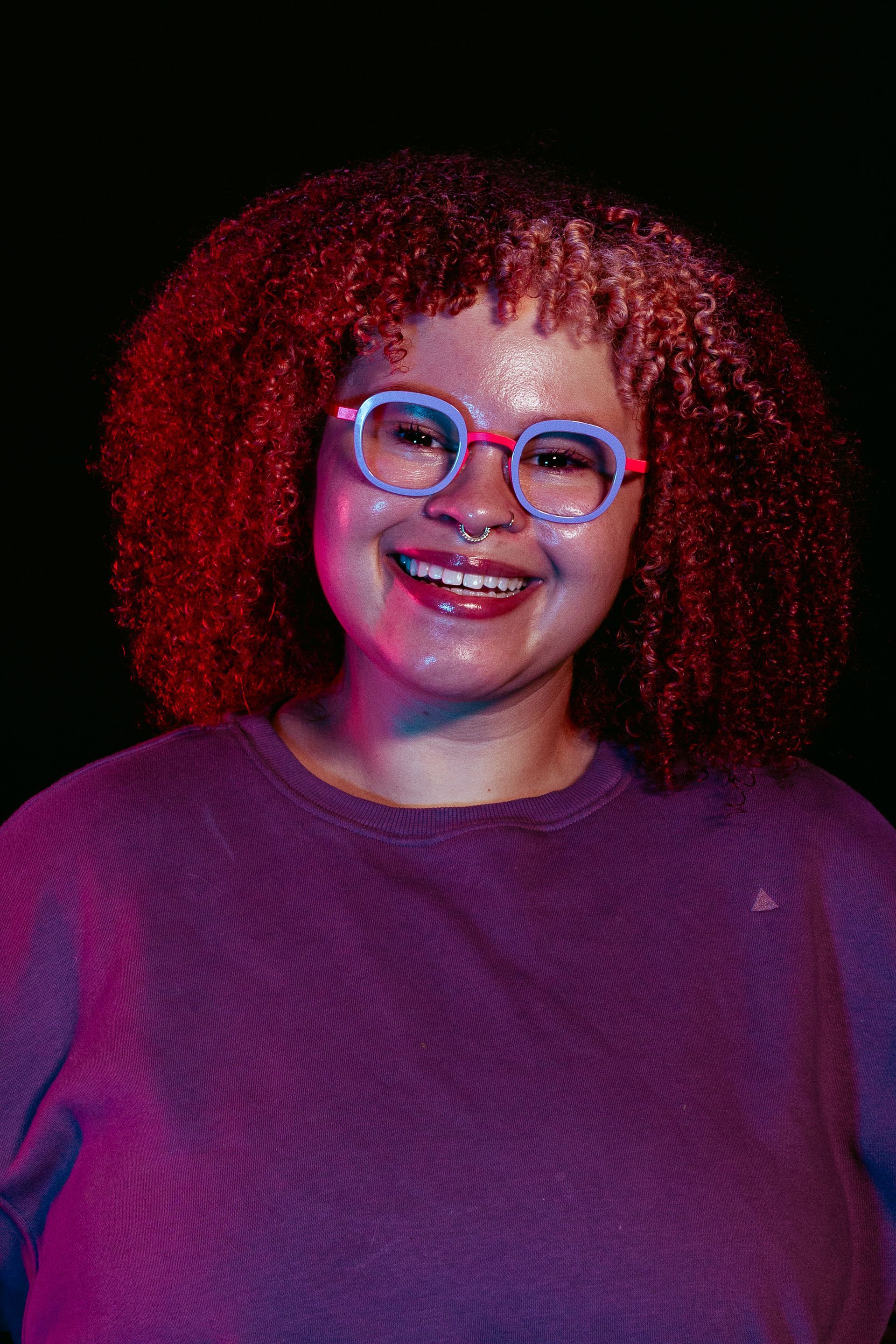 Woman with pink and red curly hair, smiling, wearing glasses and purple sweater. Black background.