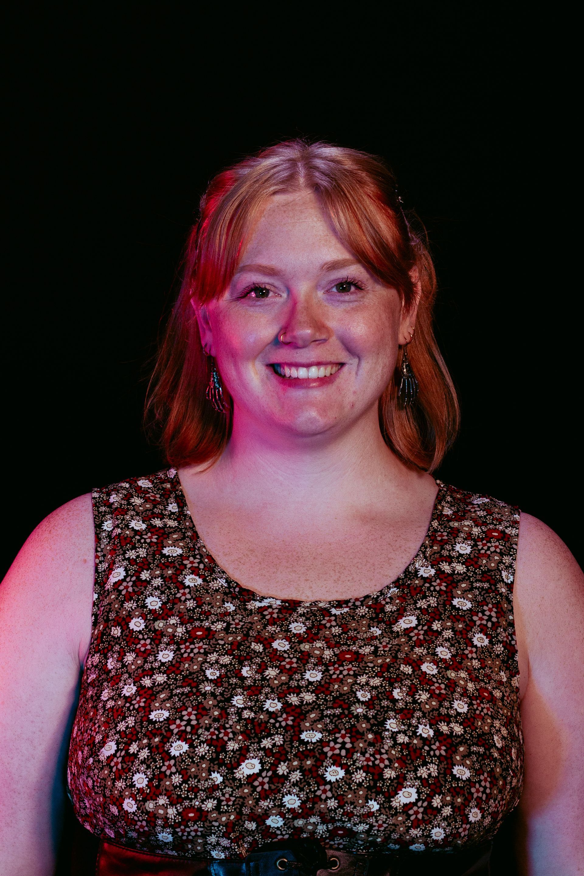 Woman with red hair and freckles smiles, wearing a floral top with red and purple lighting on a black background.