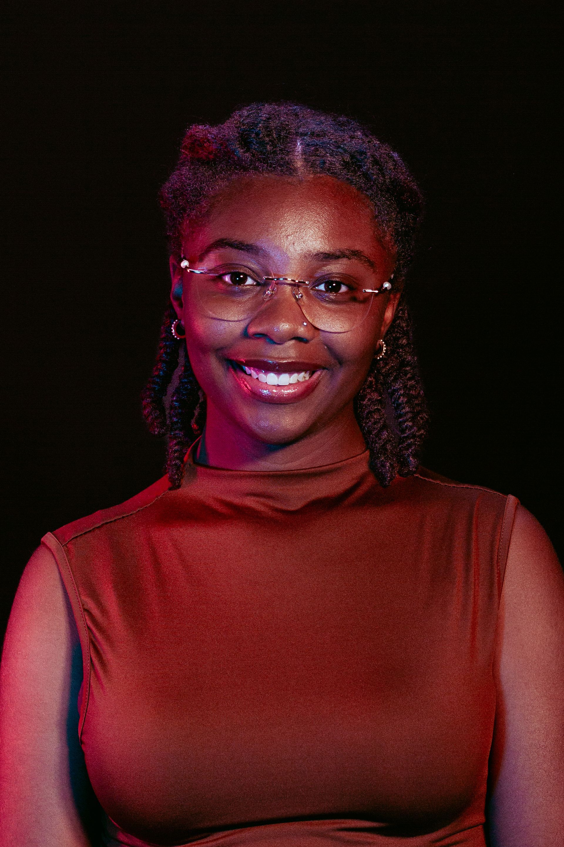 Young Black woman with glasses smiles in front of a dark backdrop, wearing a brown turtleneck.
