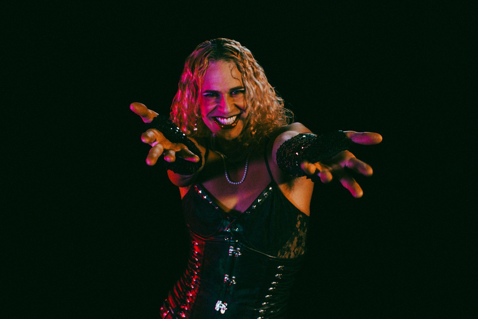 actor wearing a black corset, extending arms, smiling, in front of a dark background with red and purple lighting.