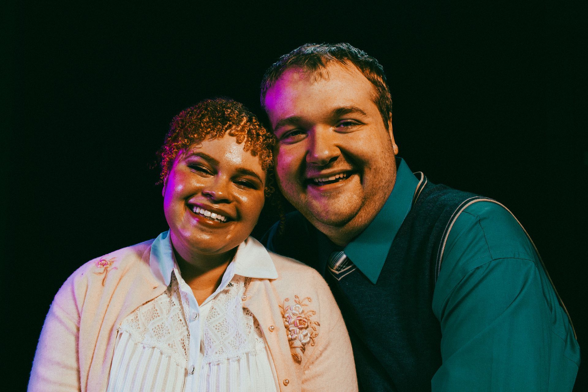 Two people smiling. Woman in pink sweater, man in blue shirt and vest, black background.