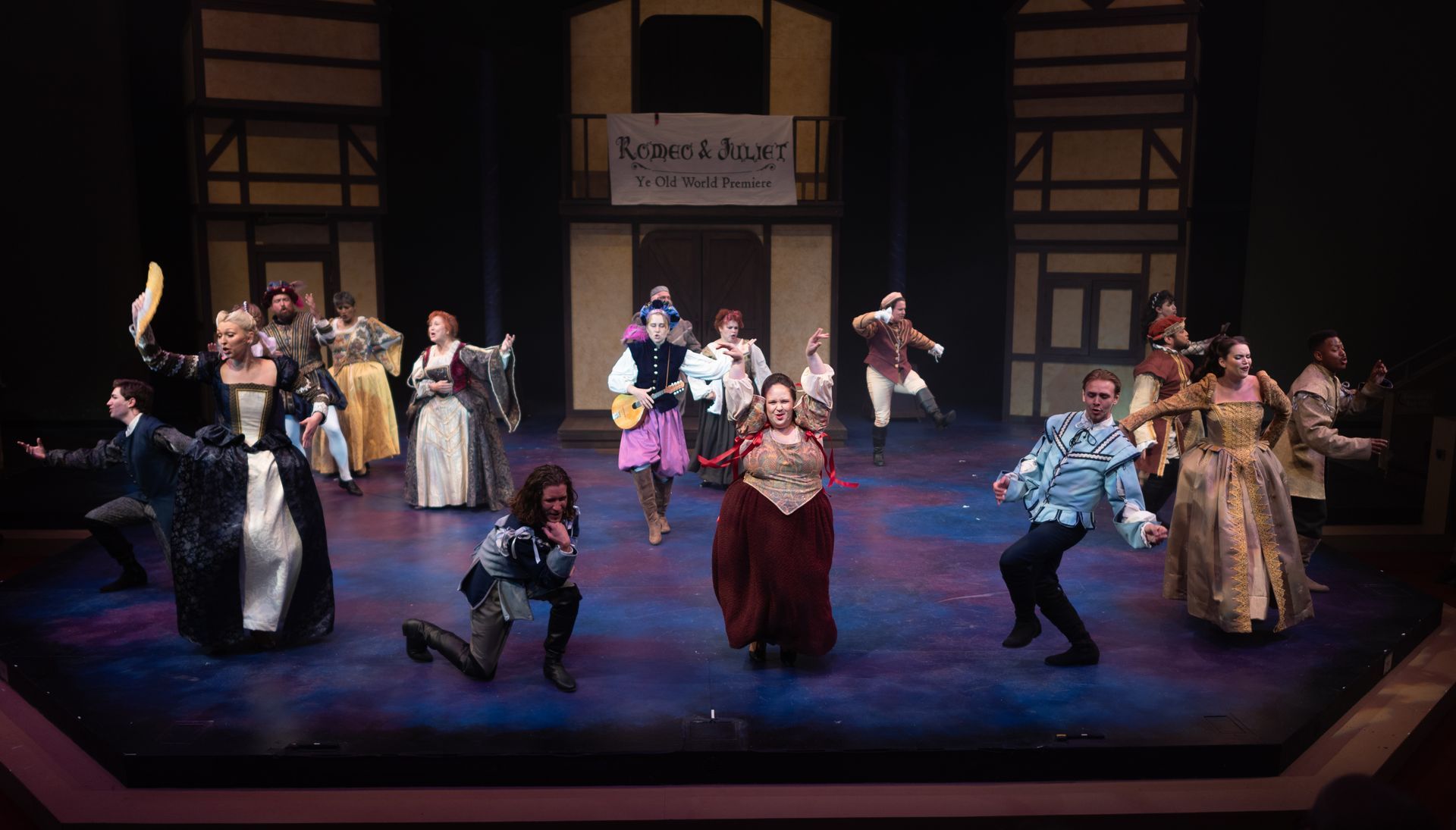 A stage play with costumed actors. One woman in red stands center, surrounded by others in elaborate attire.
