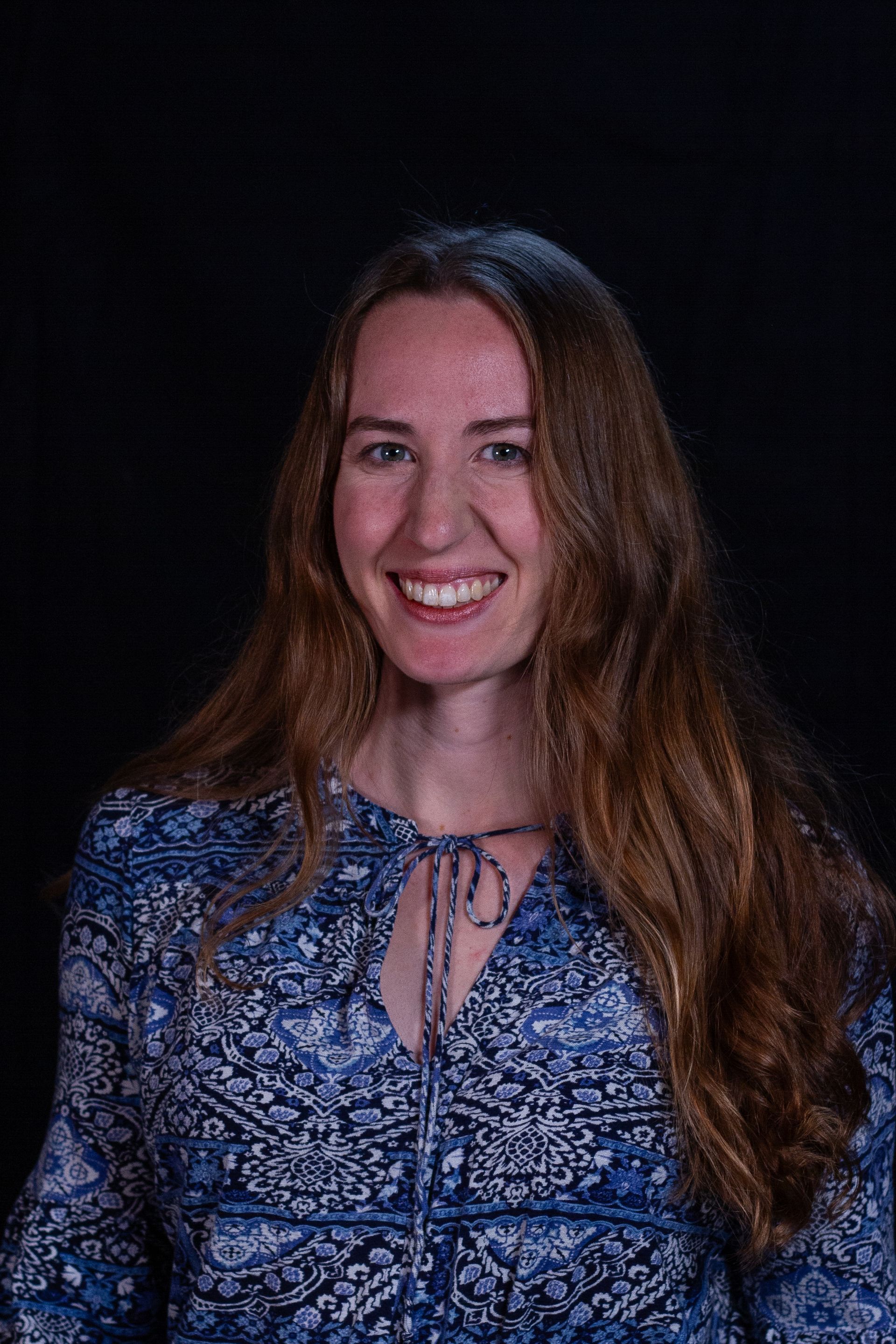 Woman with long brown hair, wearing a blue patterned top, smiling at the camera against a black background.